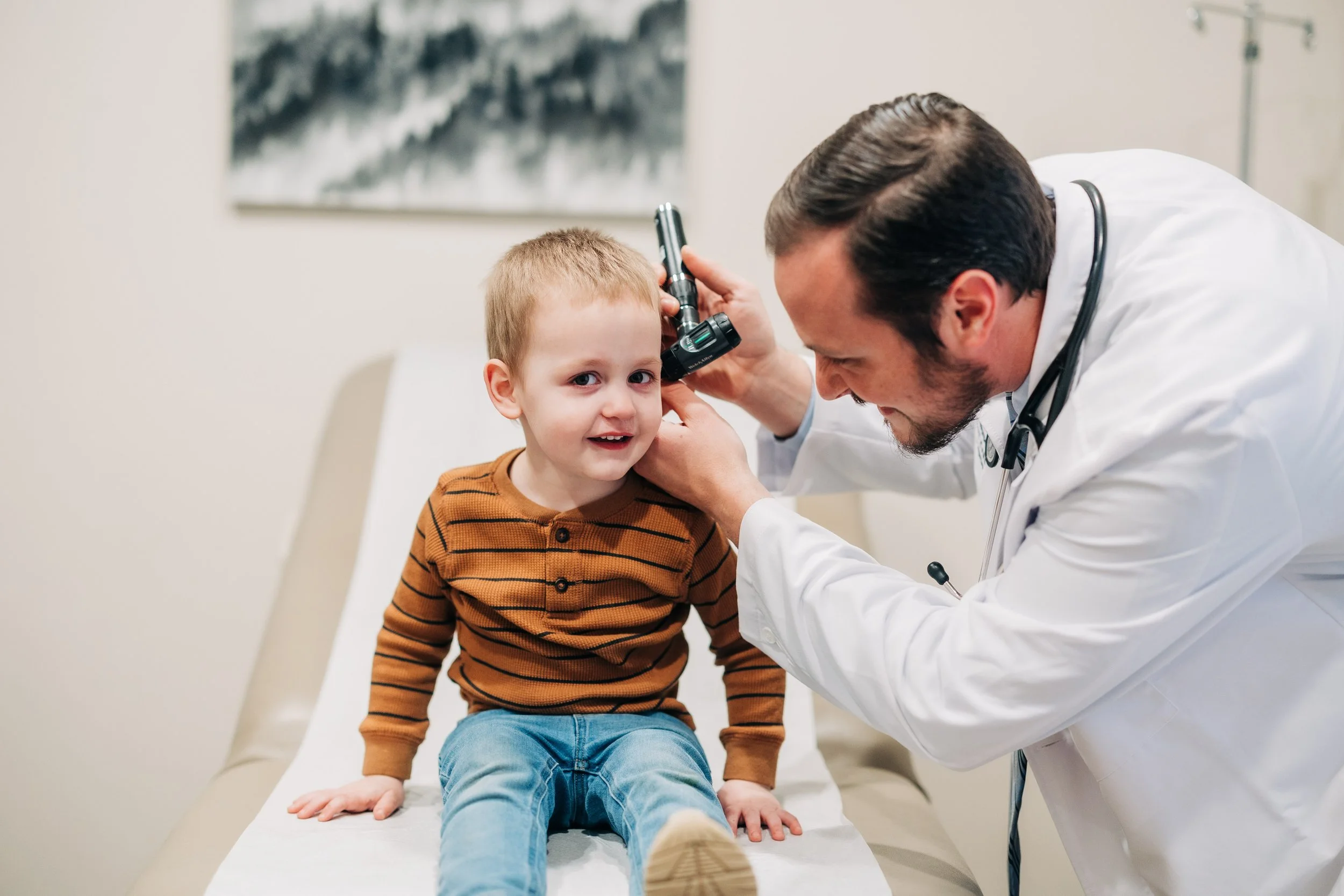 A young boy sitting on an examination table at the doctor’s office, getting his ear checked by a male doctor wearing a white coat and stethoscope, with a blurred painting on the wall in the background.