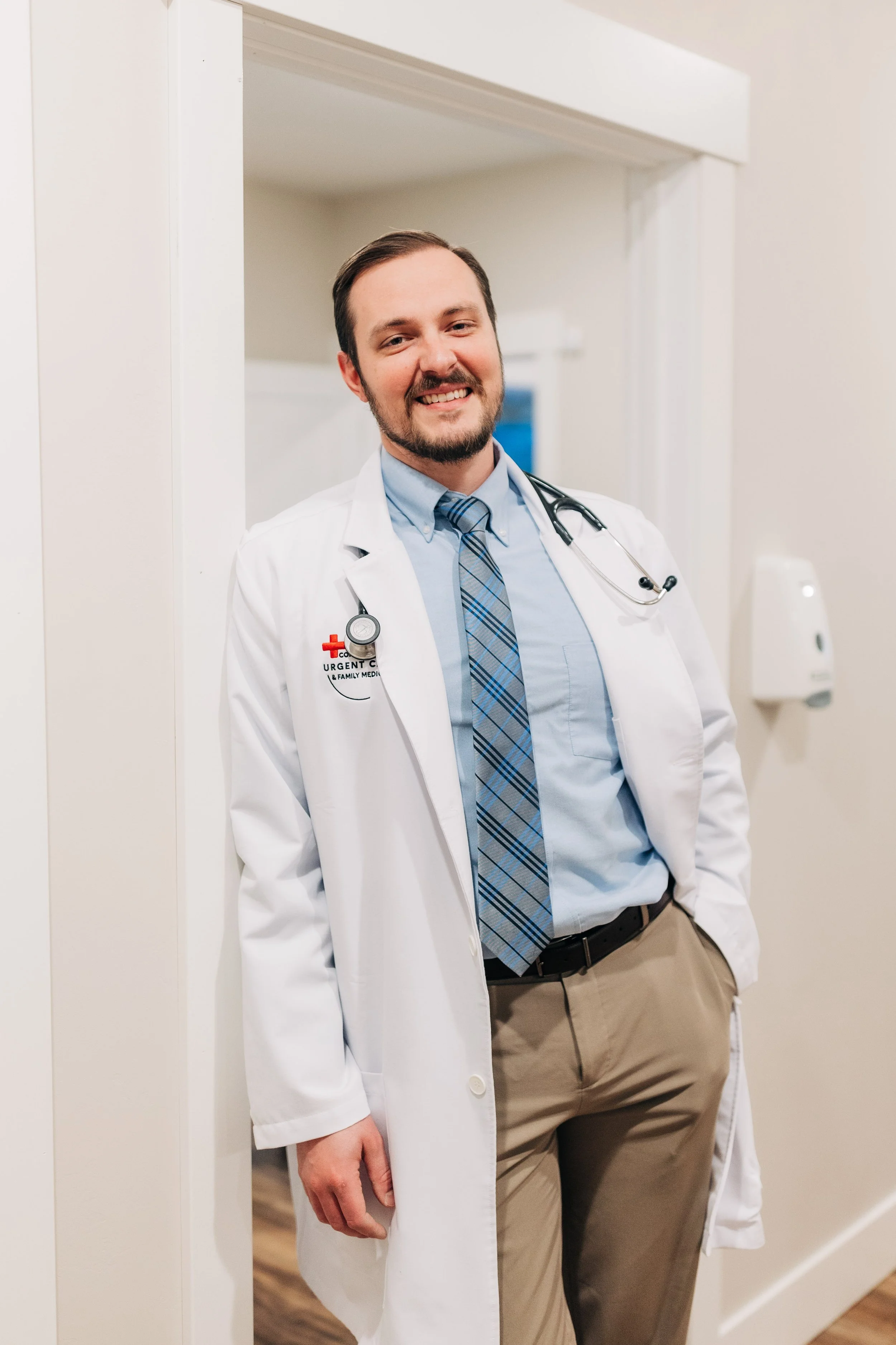 A male doctor dressed in a white coat and tie, with a stethoscope around his neck, standing in a hospital corridor, smiling at the camera.