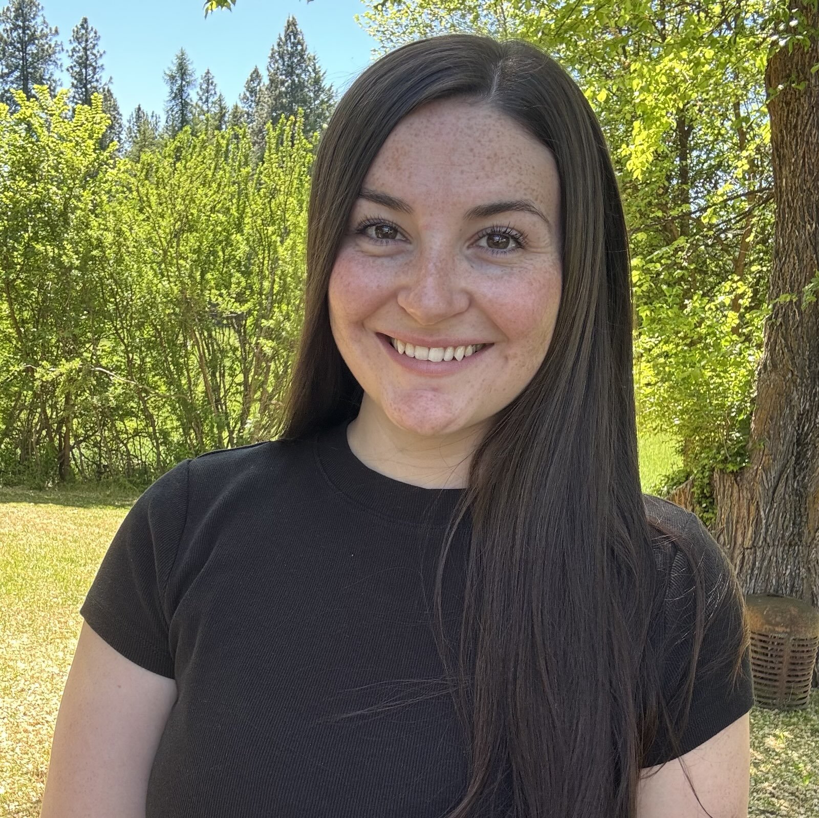 A young woman with long dark hair smiling outdoors on a sunny day, standing near trees and greenery.