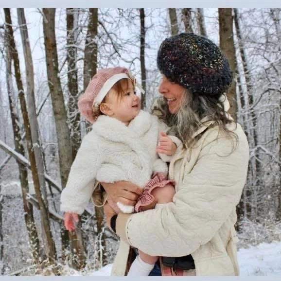 A woman and a young girl smiling and looking at each other outdoors in a snowy forest. The woman has gray hair and is wearing a black hat and a cream-colored jacket. The girl is wearing a pink hat and a white coat.