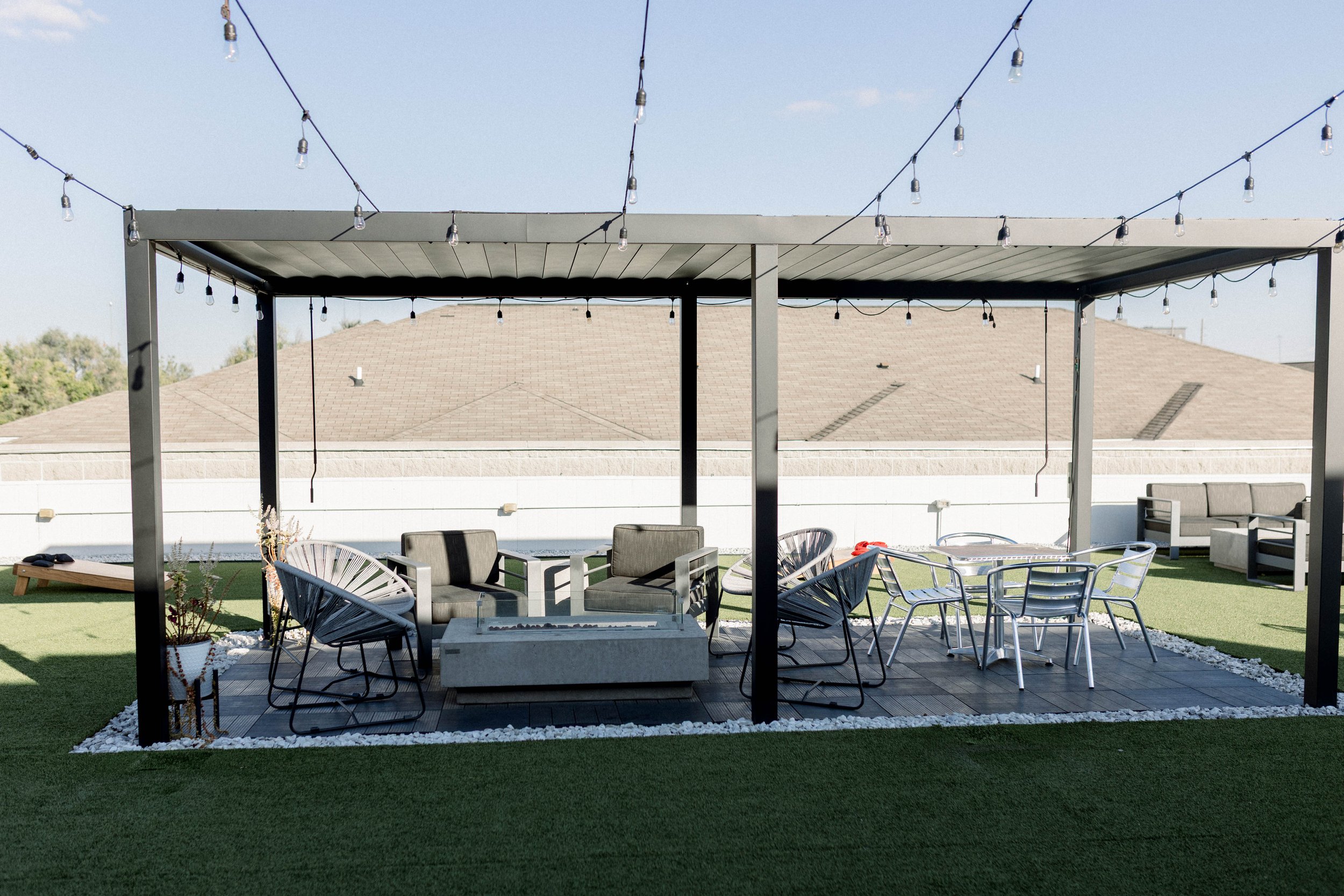 A backyard patio with outdoor furniture under a pergola with string lights, green grass, and a neighboring house roof in the background.