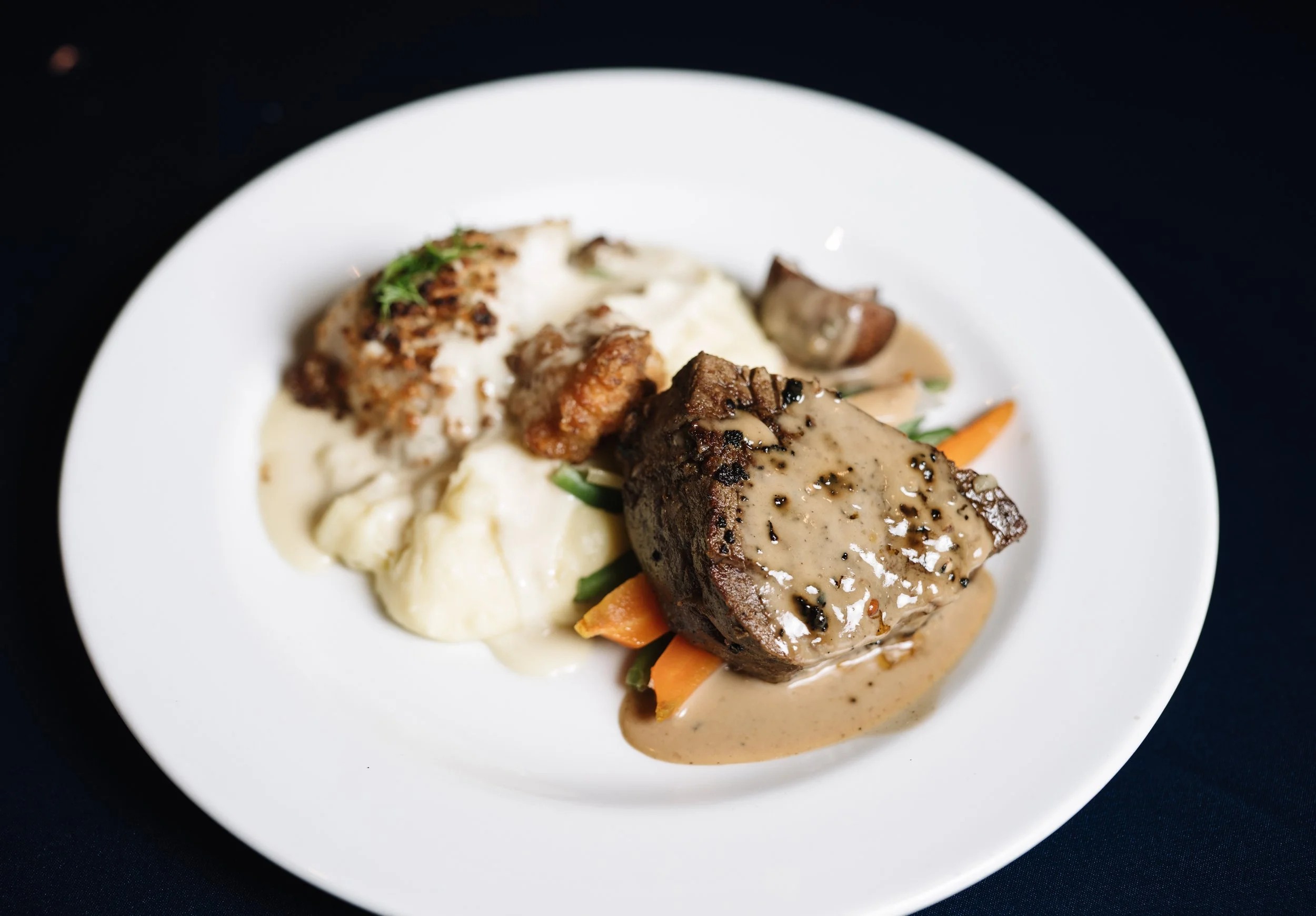 A white plate with mashed potatoes topped with herbs, cooked vegetables, and two pieces of meat smothered in a creamy gravy with black pepper, all set against a black background.