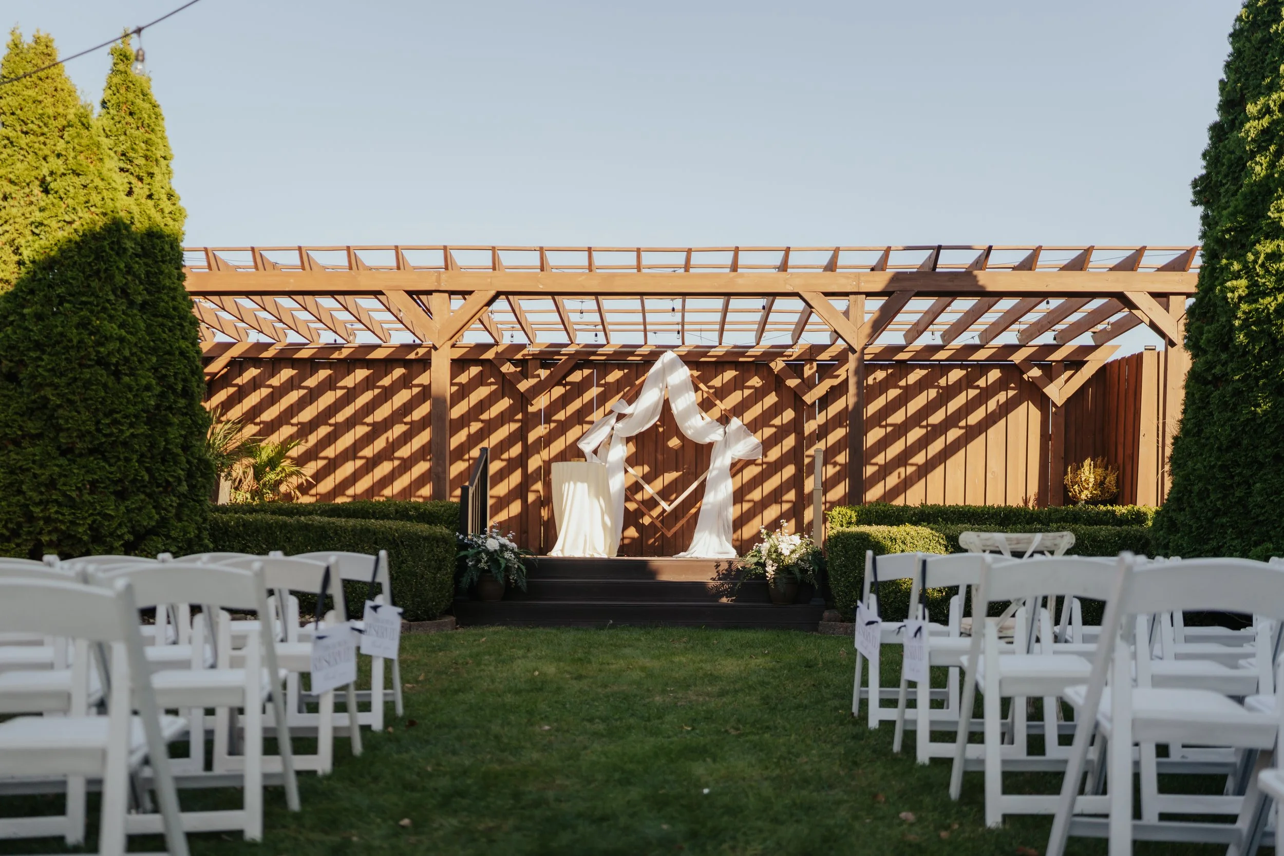 Outdoor wedding ceremony setup with white chairs facing a small stage decorated with white drapery, in front of a wooden structure with a heart-shaped decoration, flanked by tall green trees on either side.