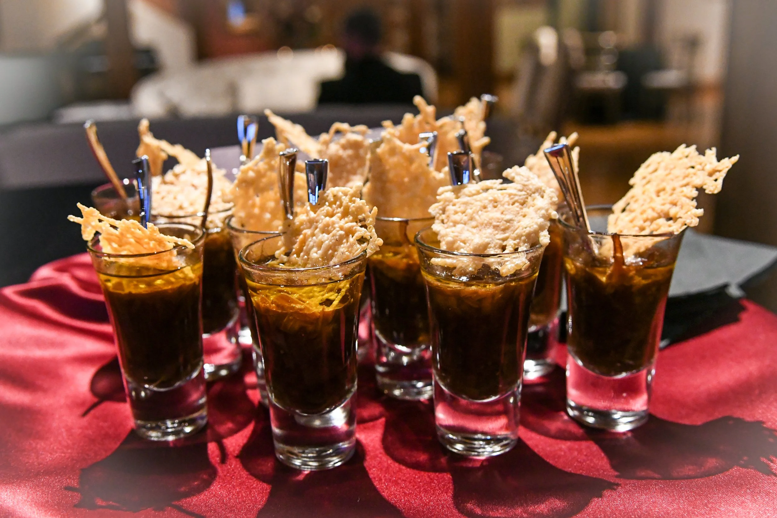 Several shot glasses filled with a dark-colored drink, topped with crispy, fried cheese snacks, arranged on a red cloth on a table.