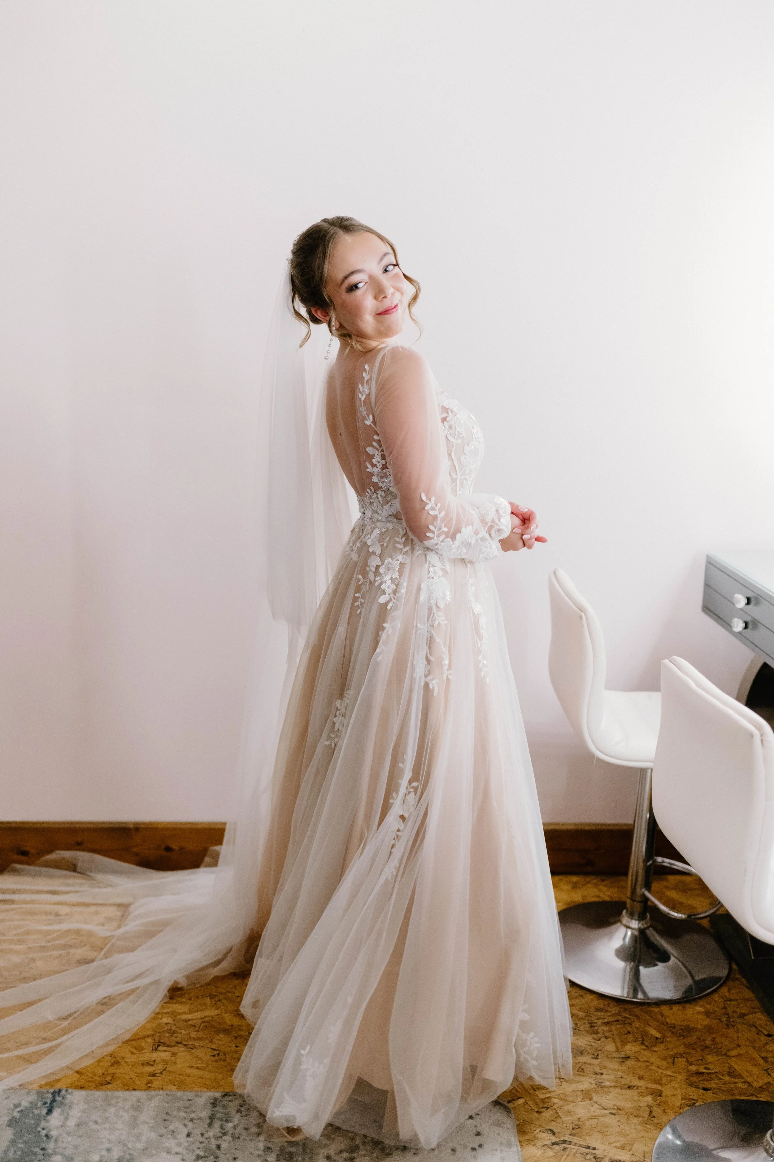 A bride in a wedding dress with lace and floral embroidery, standing indoors near a white wall, with a veil, long flowing skirt, and her hands clasped together.