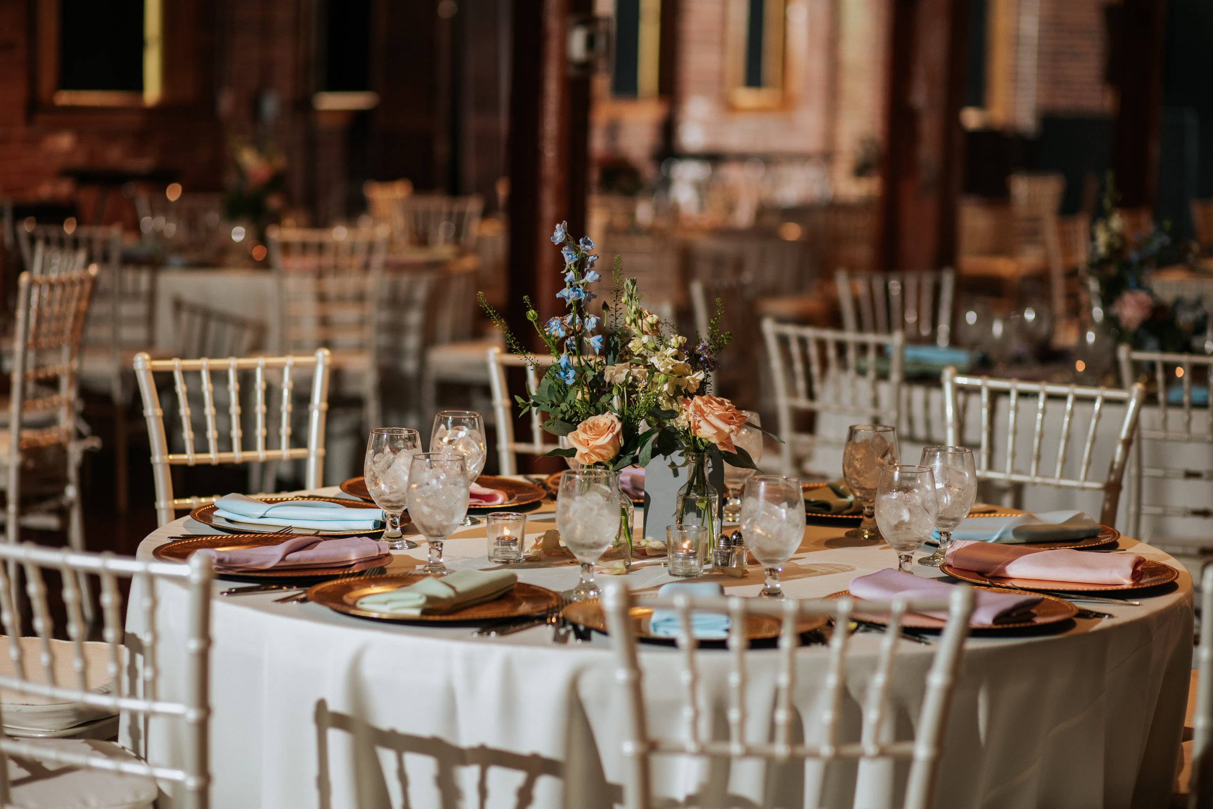 Wedding reception table with a floral centerpiece, glasses, cloth napkins, and chairs in a rustic venue with brick walls and wooden beams.