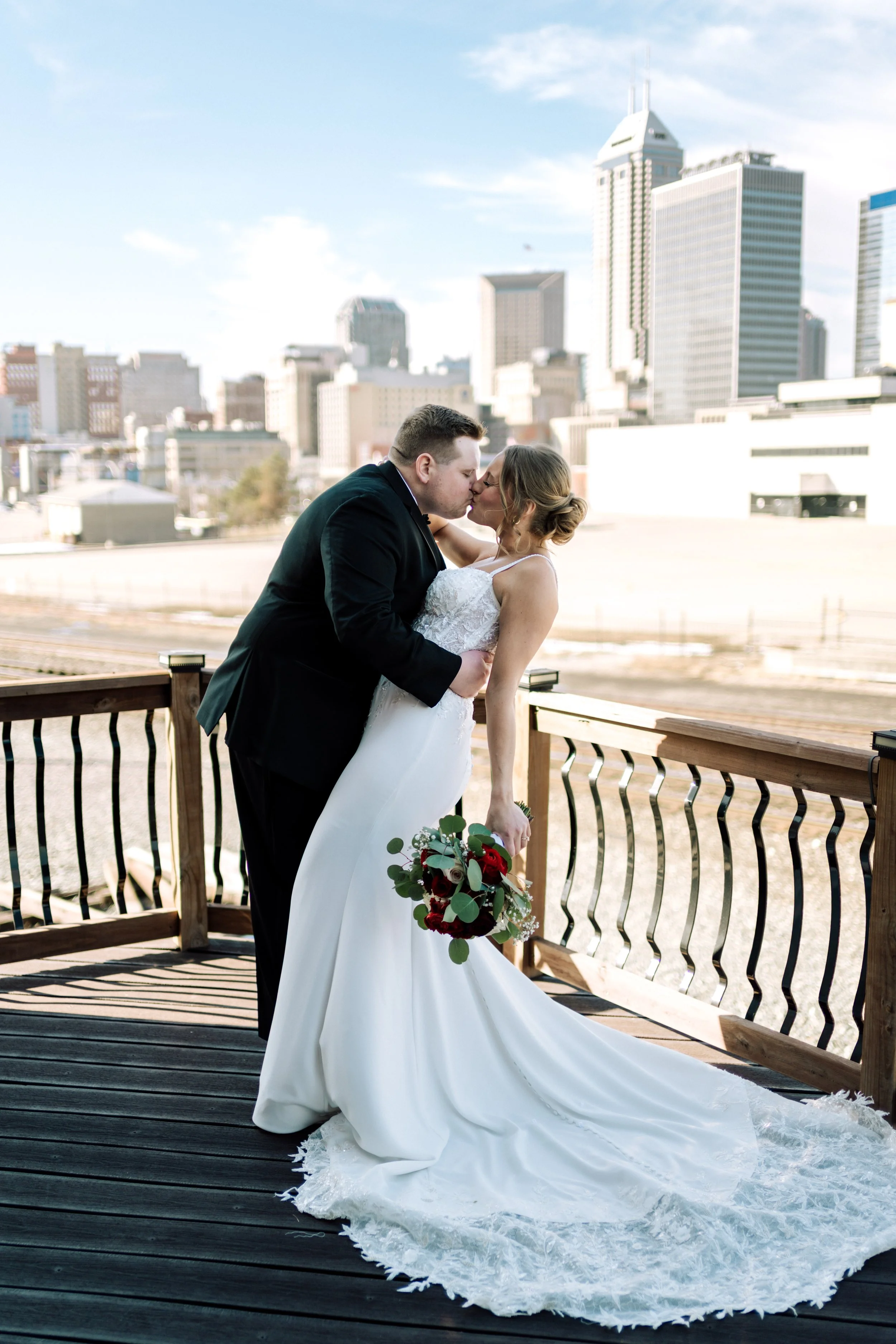 A bride and groom kissing on a wooden balcony with city skyline in the background during daytime.