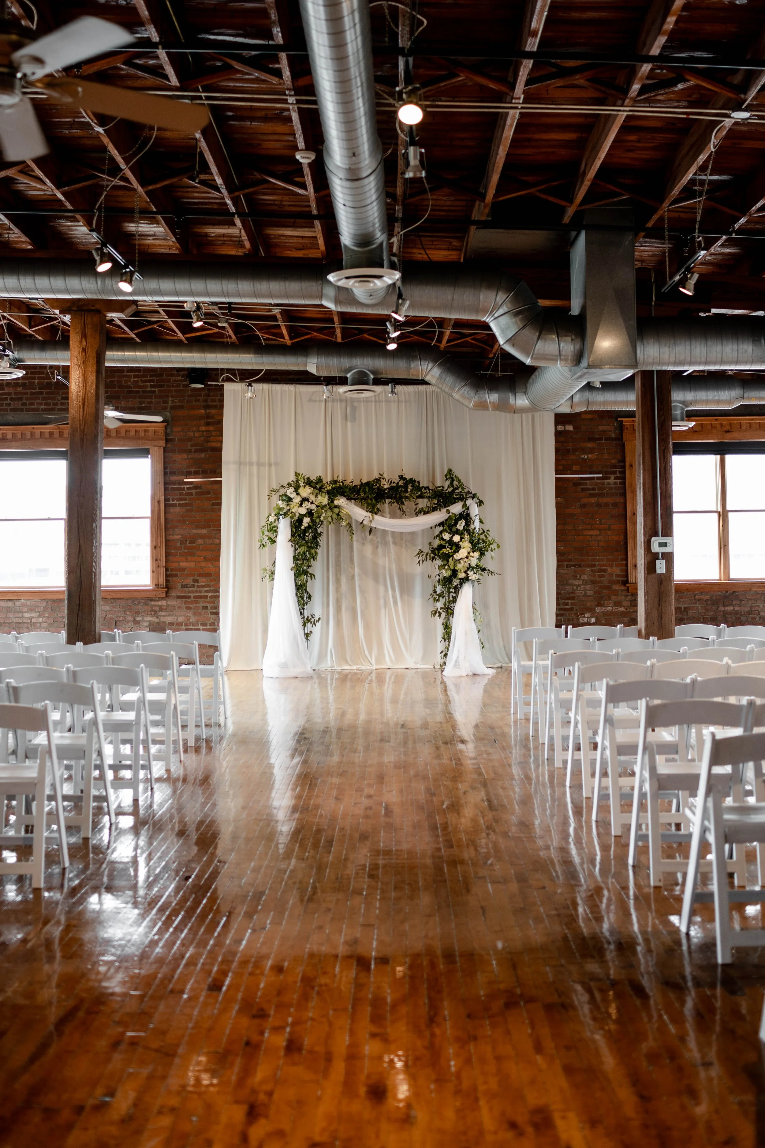 Indoor wedding ceremony setup with white chairs arranged in rows on a wooden floor, a floral arch, and a white fabric backdrop in a brick-walled industrial-style venue with exposed ductwork and large windows.