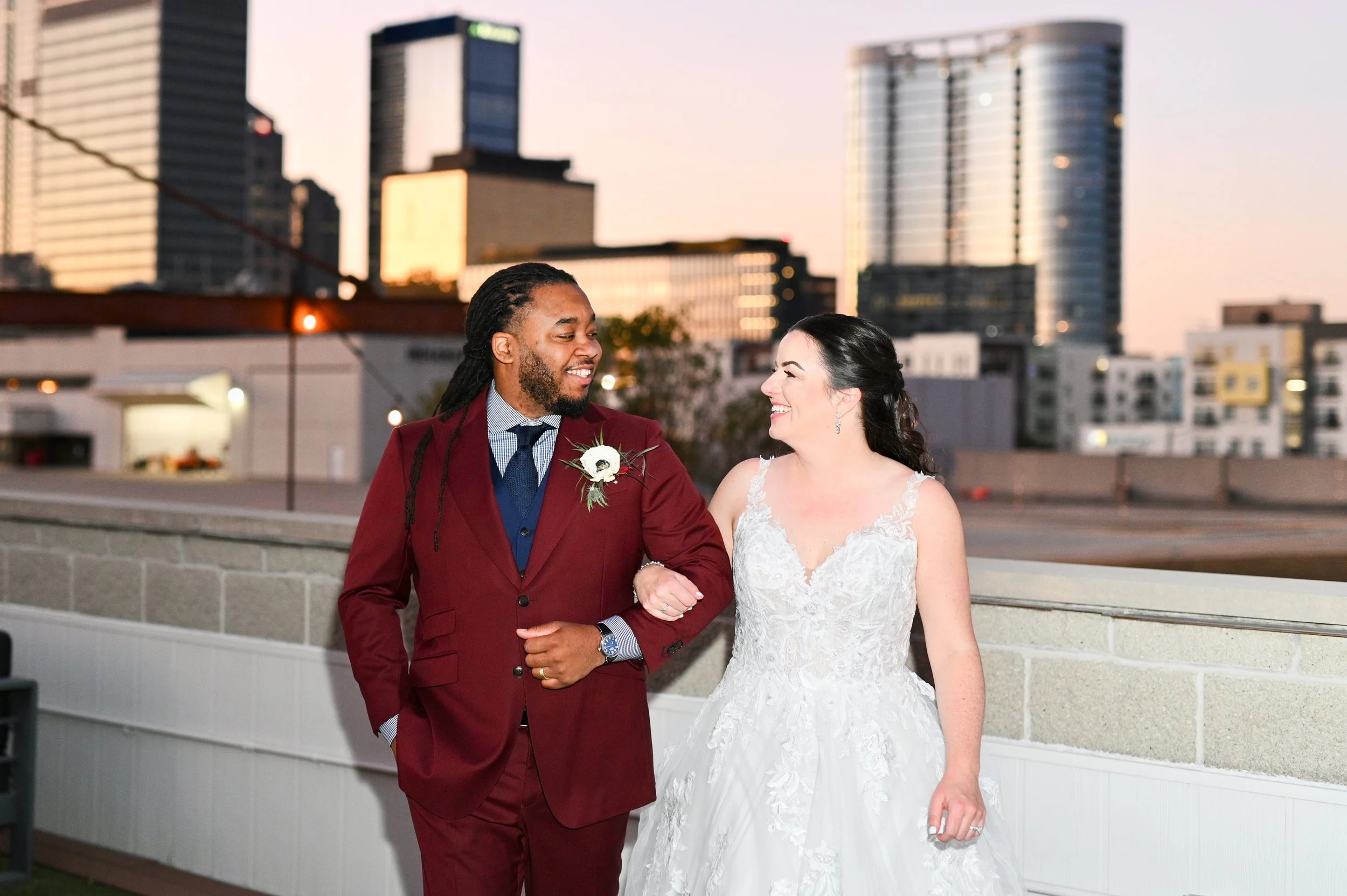A bride and groom holding hands and smiling, standing outdoors on a rooftop with city skyline in the background during sunset. The groom wears a maroon suit with a white boutonniere, and the bride wears a white lace wedding dress with dark hair style