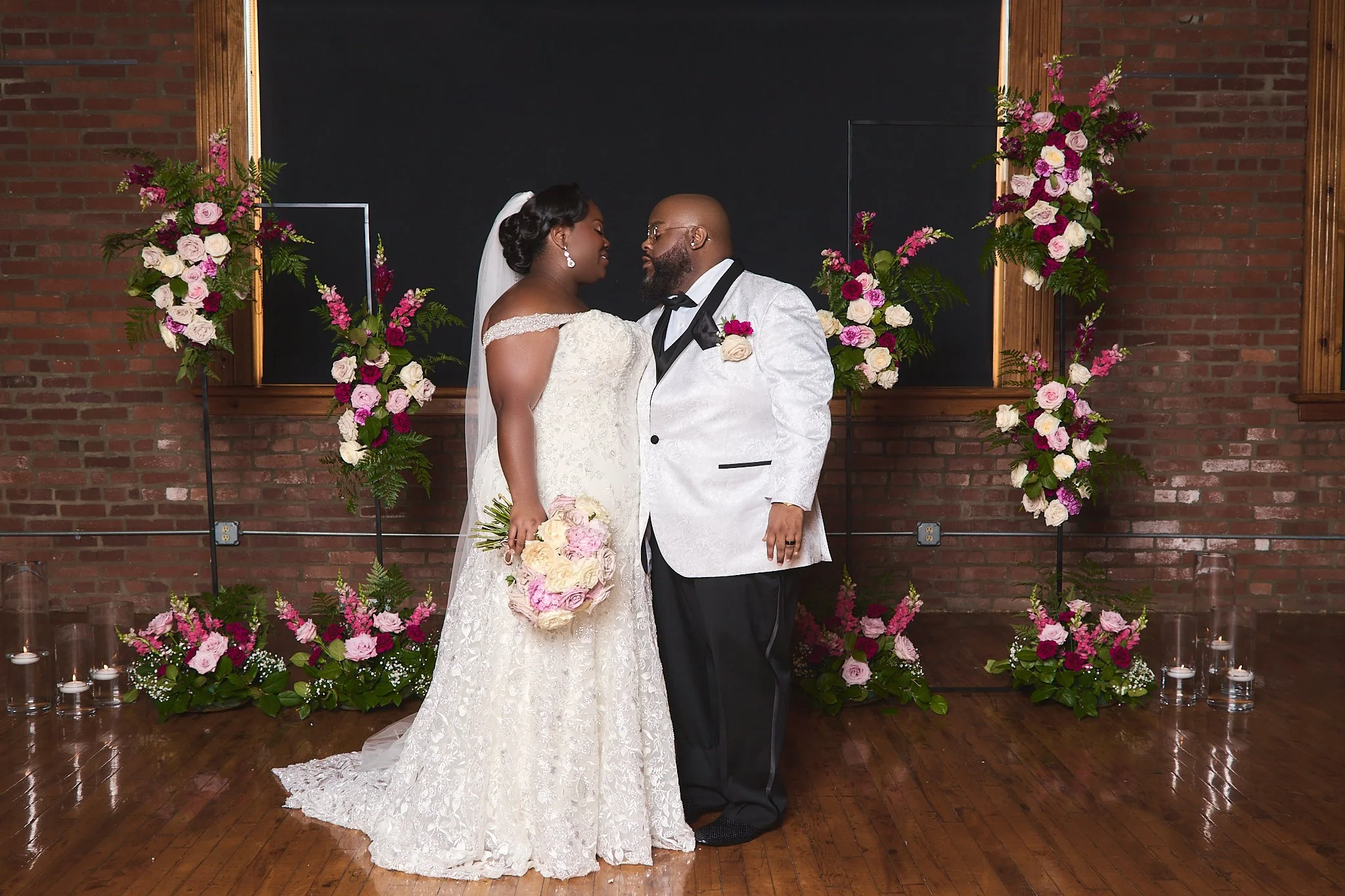 A bride and groom share a moment during their wedding ceremony, standing in front of a floral arch with pink and white flowers set against a brick wall.