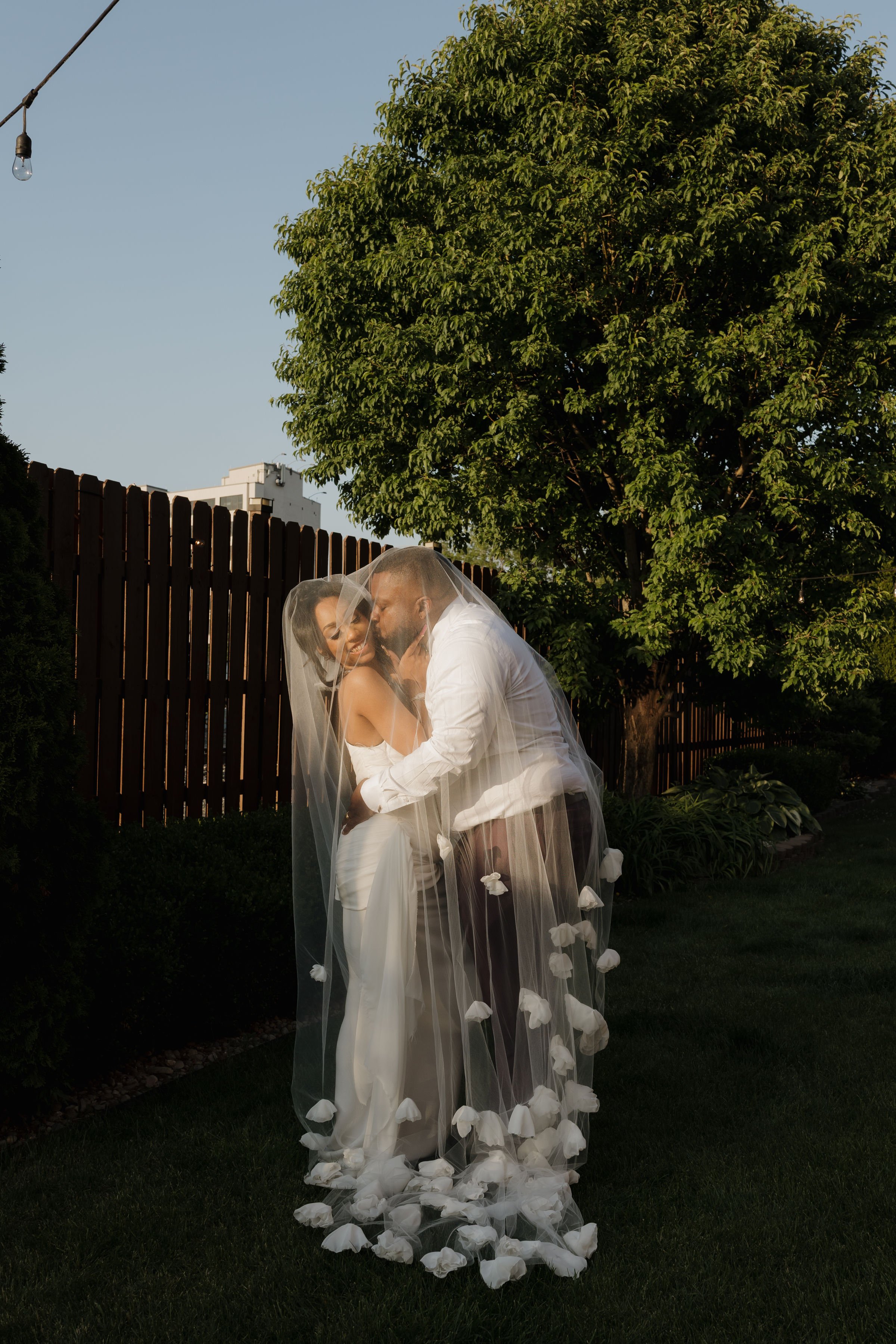 A couple dressed in wedding attire sharing an intimate moment outdoors at sunset, with a large green tree, wooden fence, and string lights in the background.