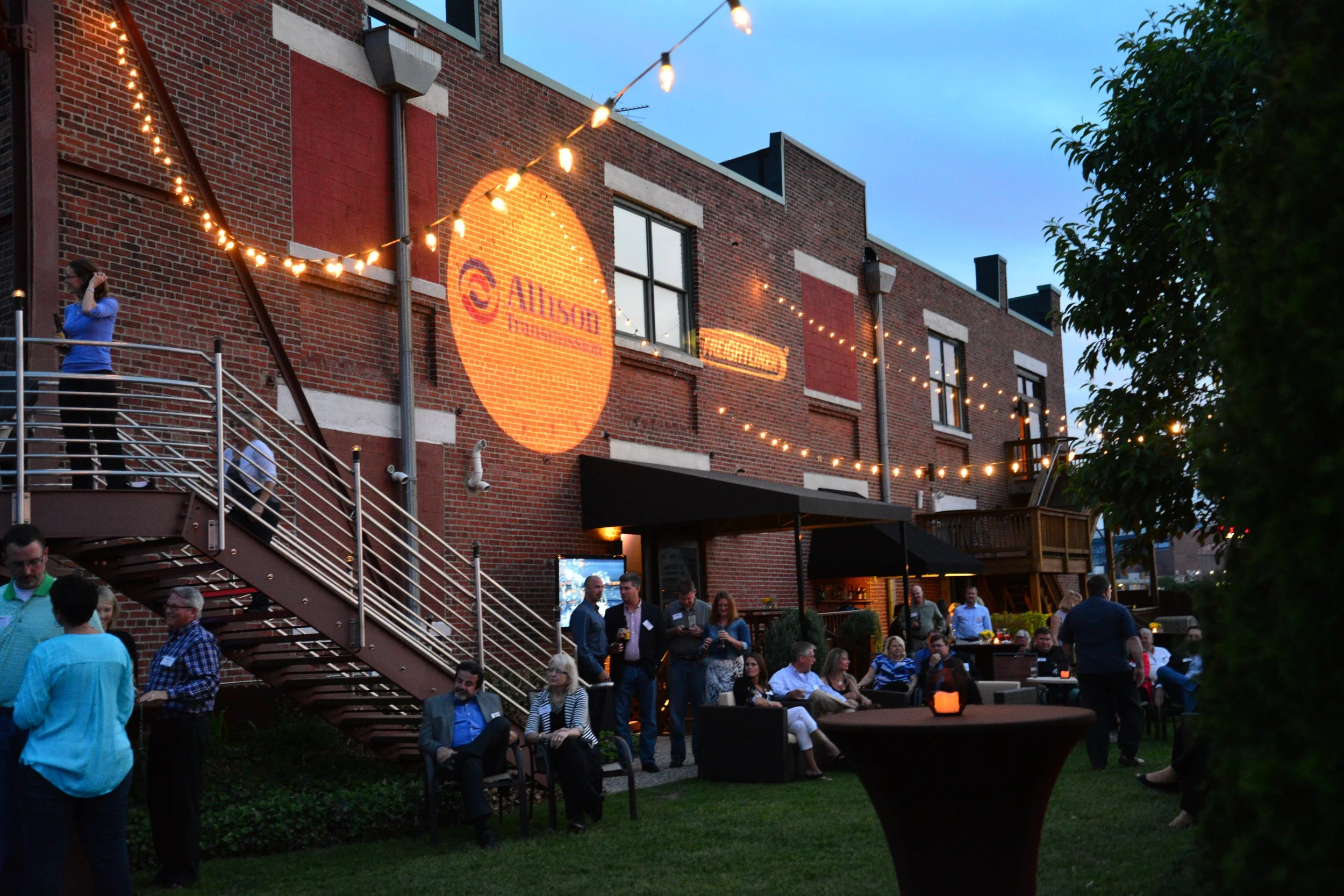 People gathered at an outdoor evening event in a courtyard with string lights, a brick building with a logo projected onto it, and various groups of guests socializing and sitting on chairs.