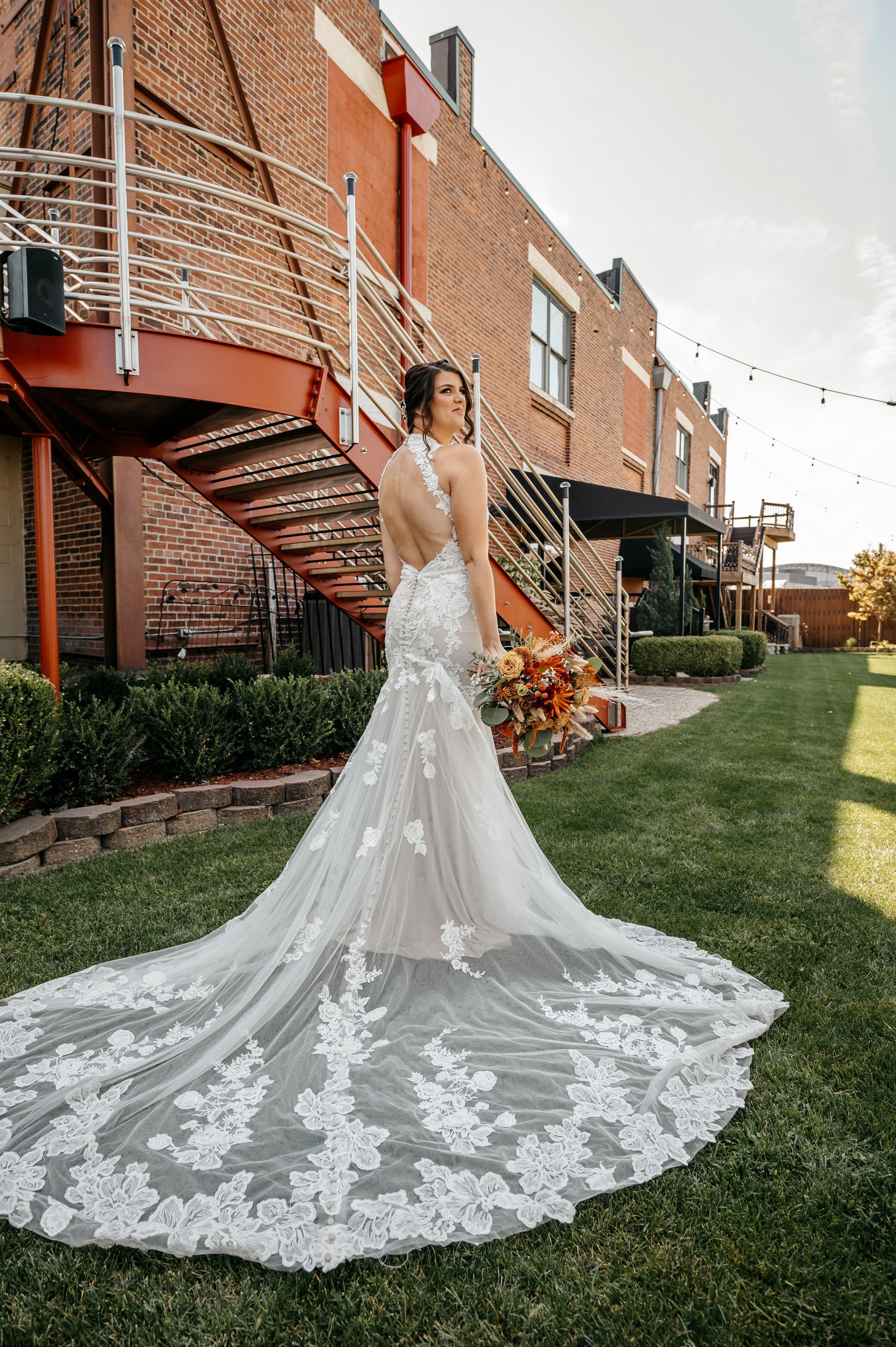 Bride in a white wedding dress with lace appliqué, holding a bouquet of flowers, standing outdoors on a grassy lawn near a brick building with a spiral staircase.