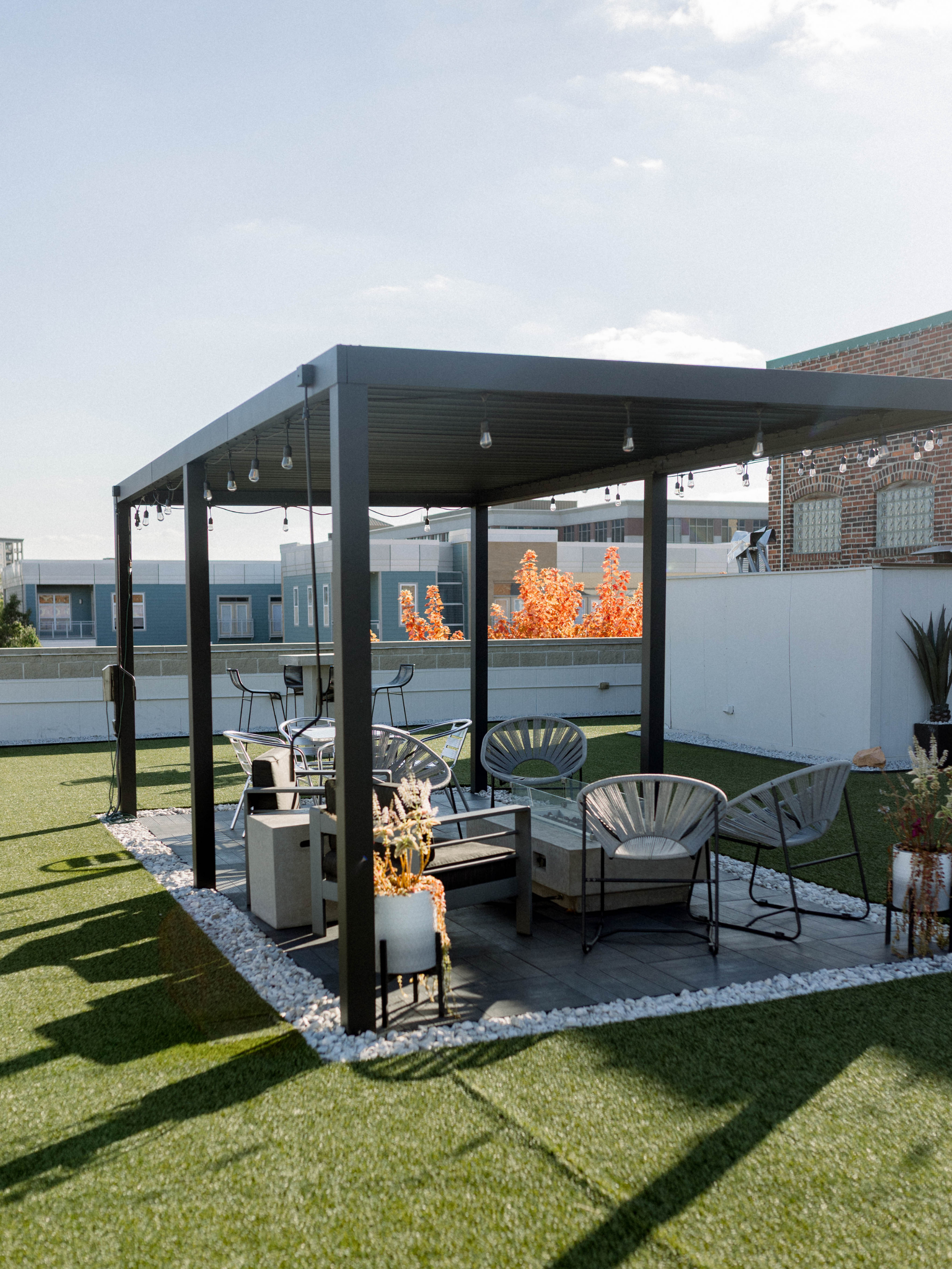 Outdoor patio with black pergola, patio furniture including chairs and a coffee table, surrounded by artificial grass and white rocks, with a brick building and trees with autumn foliage in the background under a blue sky.