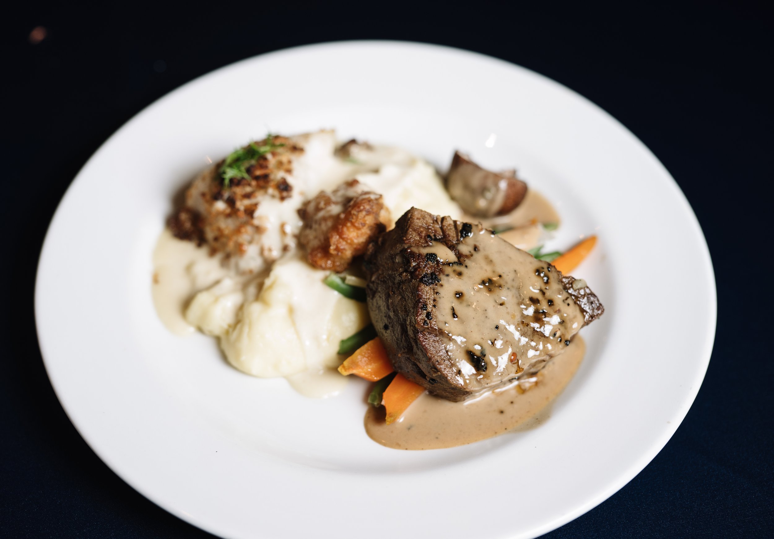 A white plate with mashed potatoes topped with herbs and mashed meatloaf with brown gravy, and a vegetable garnish including carrots and green beans, placed on a dark surface.