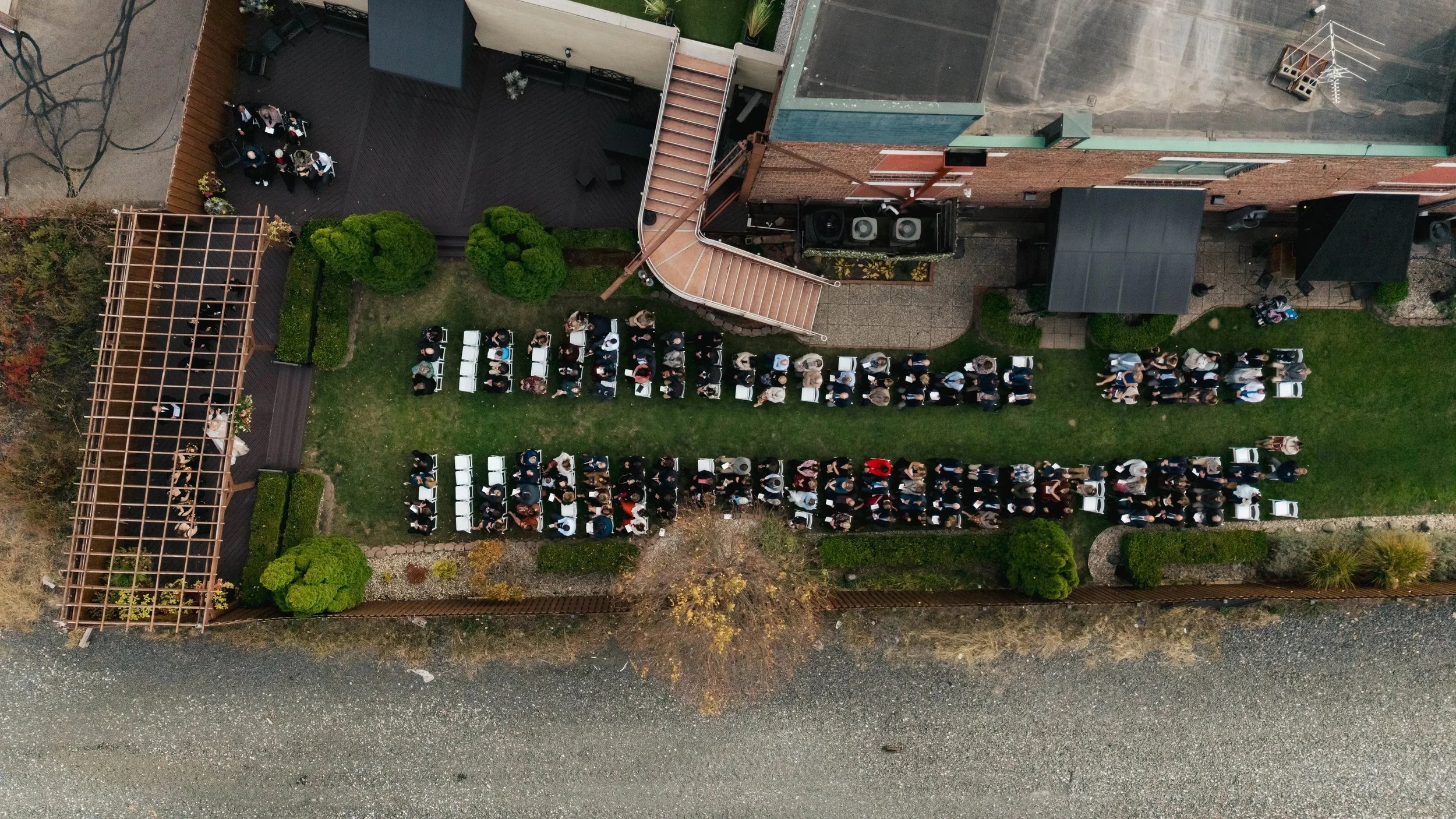 An aerial view of a backyard with a gathering of people. There are rows of chairs set up, some with guests seated, and some standing around. The yard features a wooden deck, a small garden, and a structure with a pergola. There are trees and shrubs a