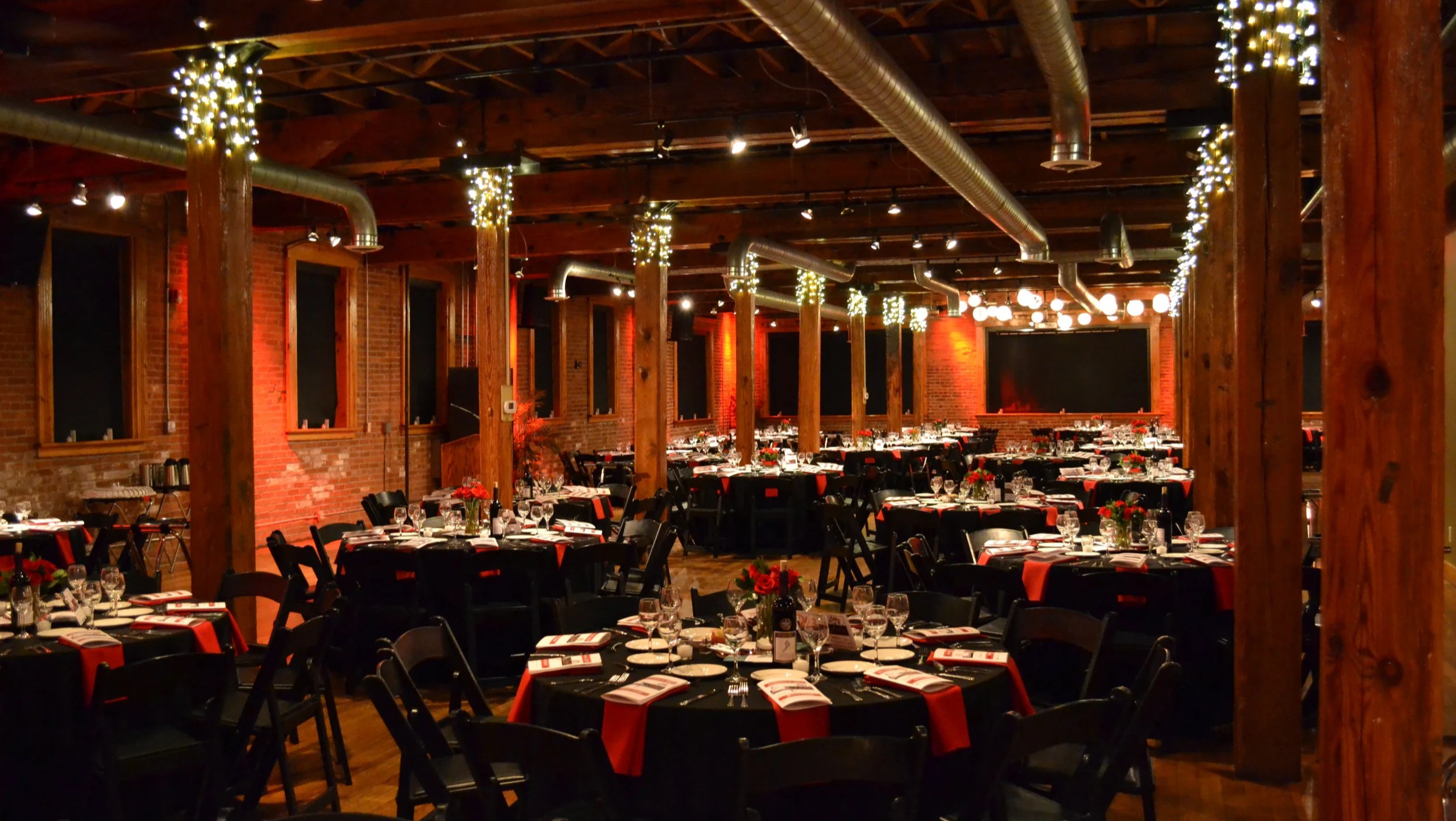 A decorated indoor event space with brick walls, wooden beams, string lights, and multiple tables set with glassware and red flowers for a formal dinner or celebration.