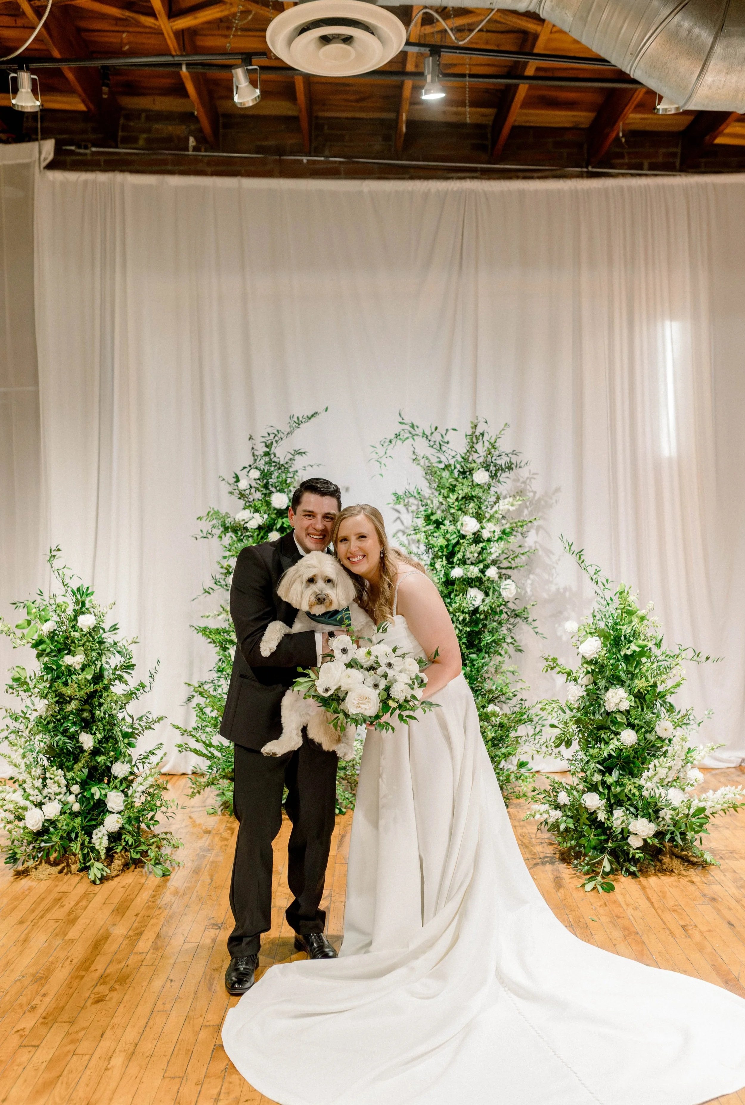 A bride and groom smiling at a wedding with a white dog, standing in front of white curtains and floral arrangements.