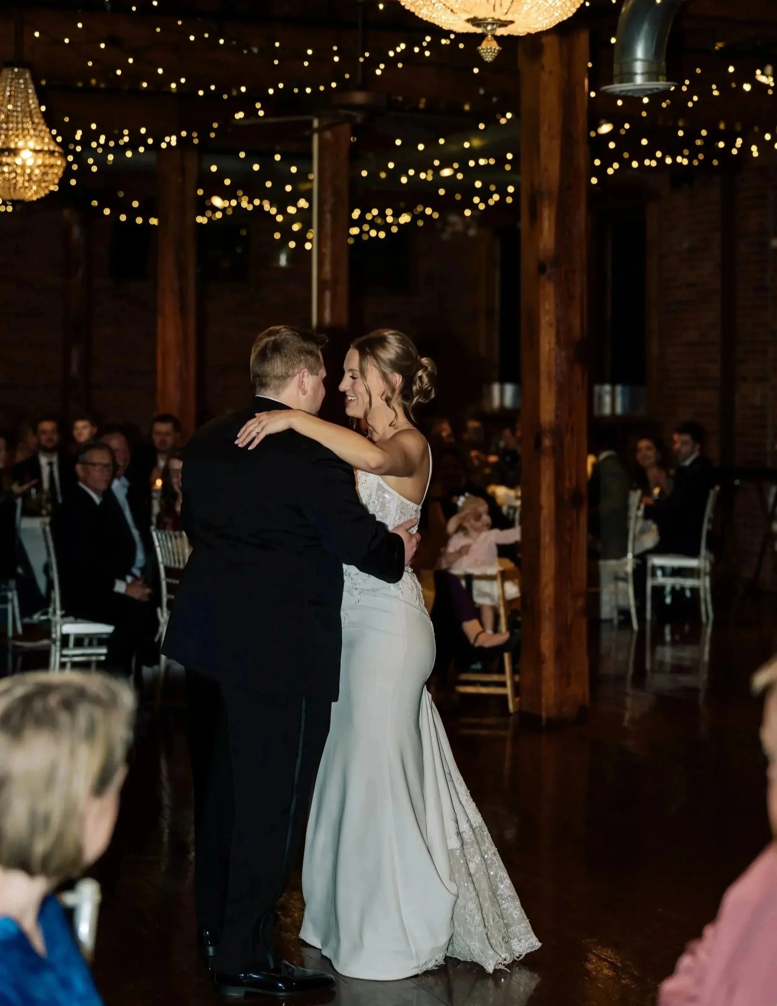 A bride and groom are dancing together at their wedding reception, inside a rustic venue with wooden beams and hanging string lights and chandeliers.