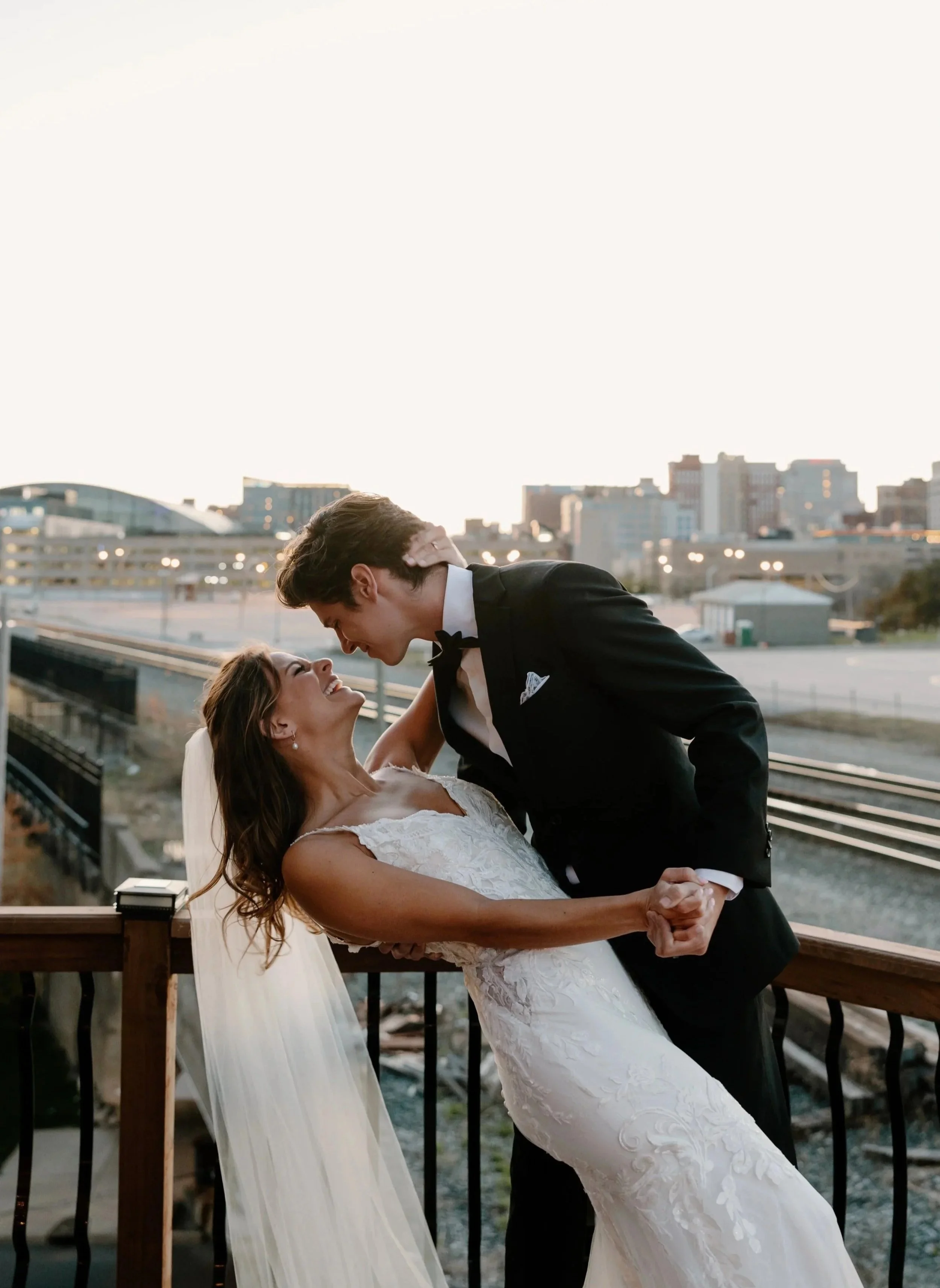 A bride and groom dancing on a balcony in an urban setting during sunset, smiling and looking at each other.