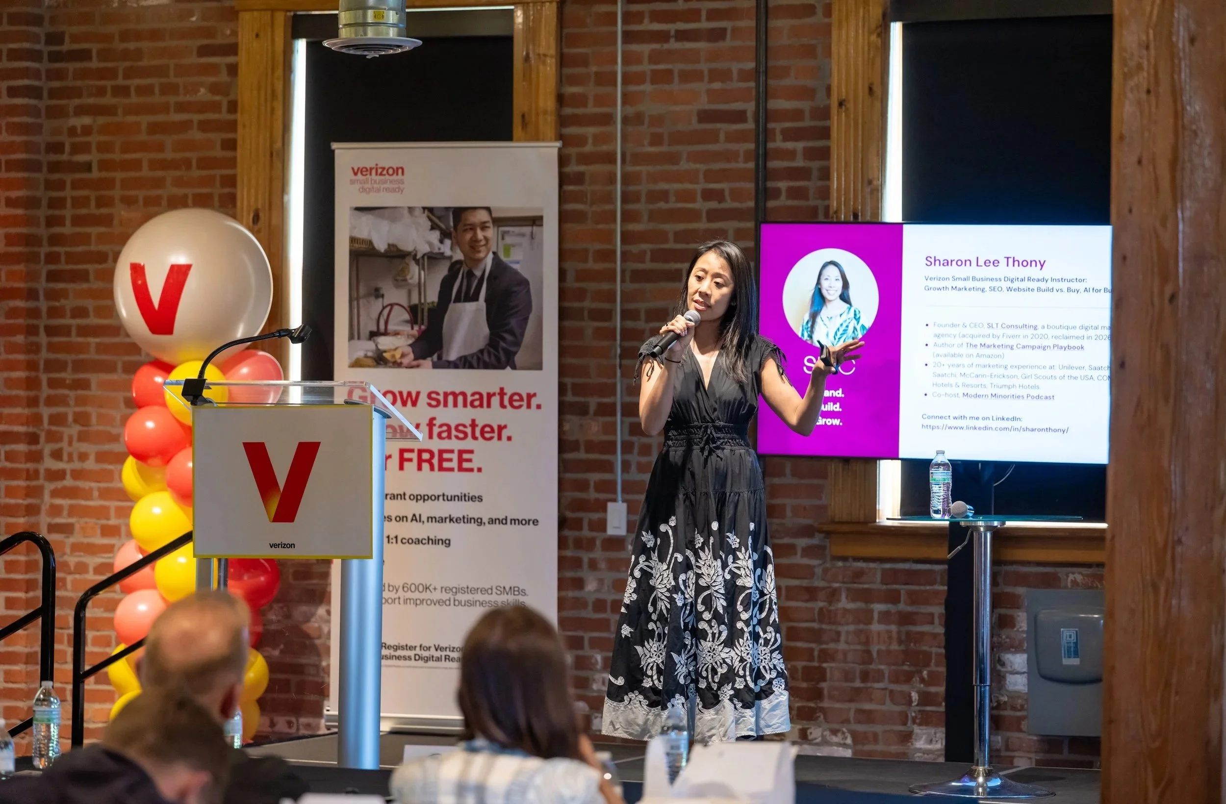 A woman giving a presentation at a conference with a large screen displaying her name, title, and information. The event features Verizon branding, with balloons and a brick wall background.