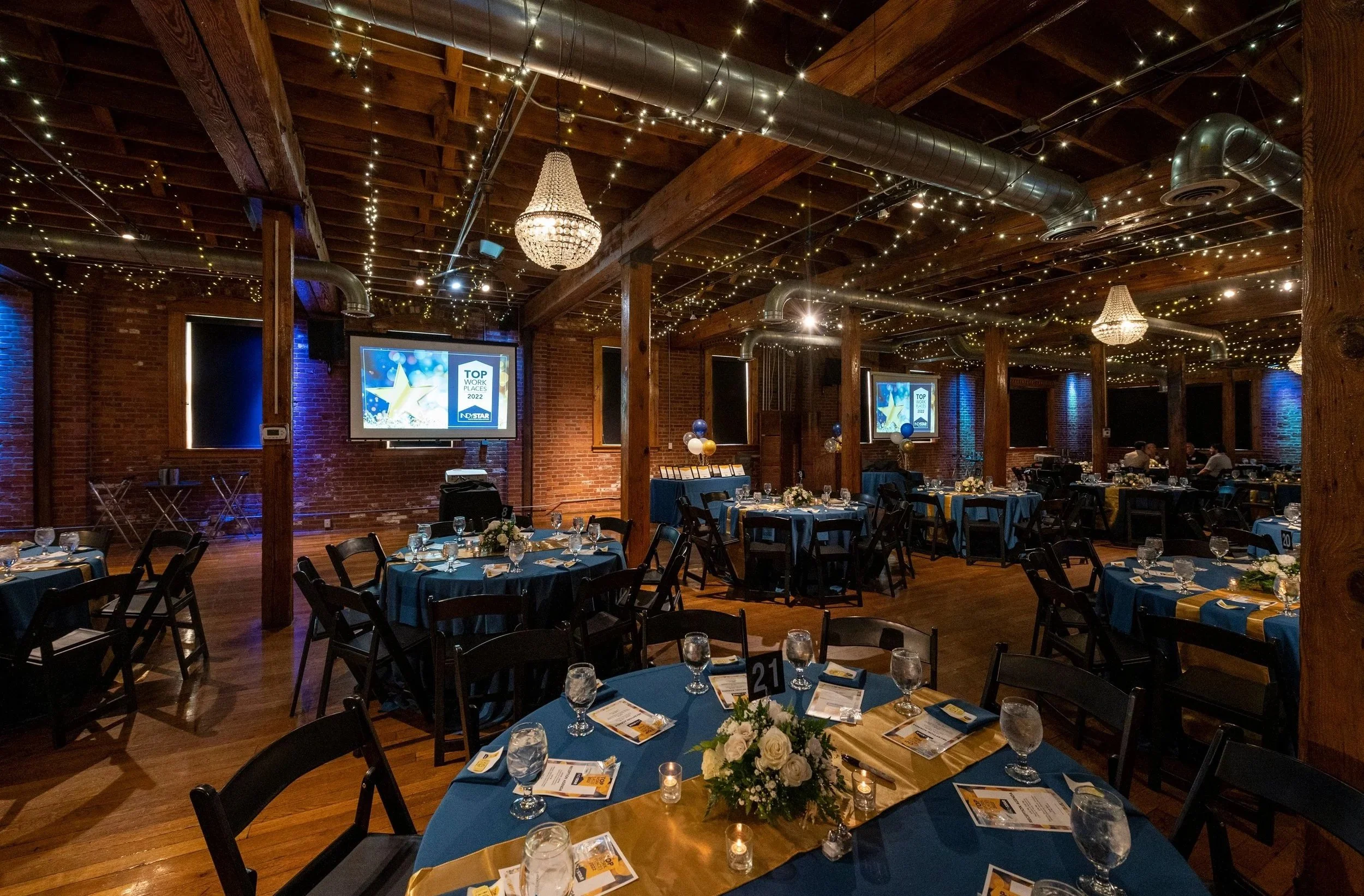 A banquet hall decorated with string lights, chandeliers, and tents with tables set for an event, featuring floral centerpieces, candles, and place settings.