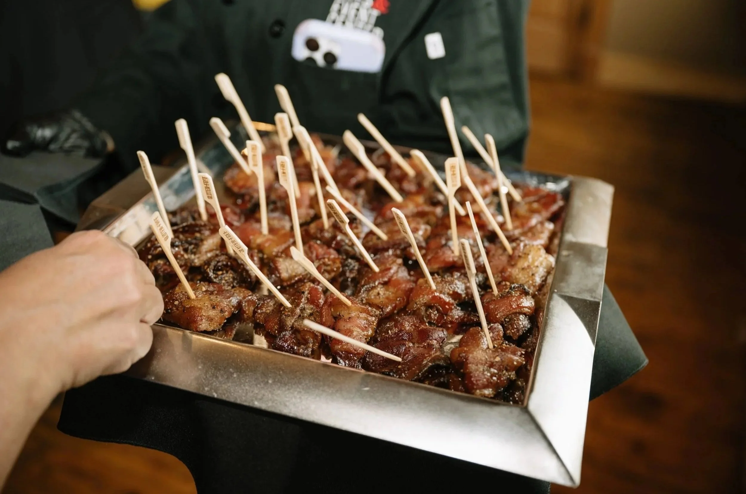 A tray of cooked meat skewers with small wooden sticks inserted into each piece, ready for serving or eating, with a person's hand reaching for one.