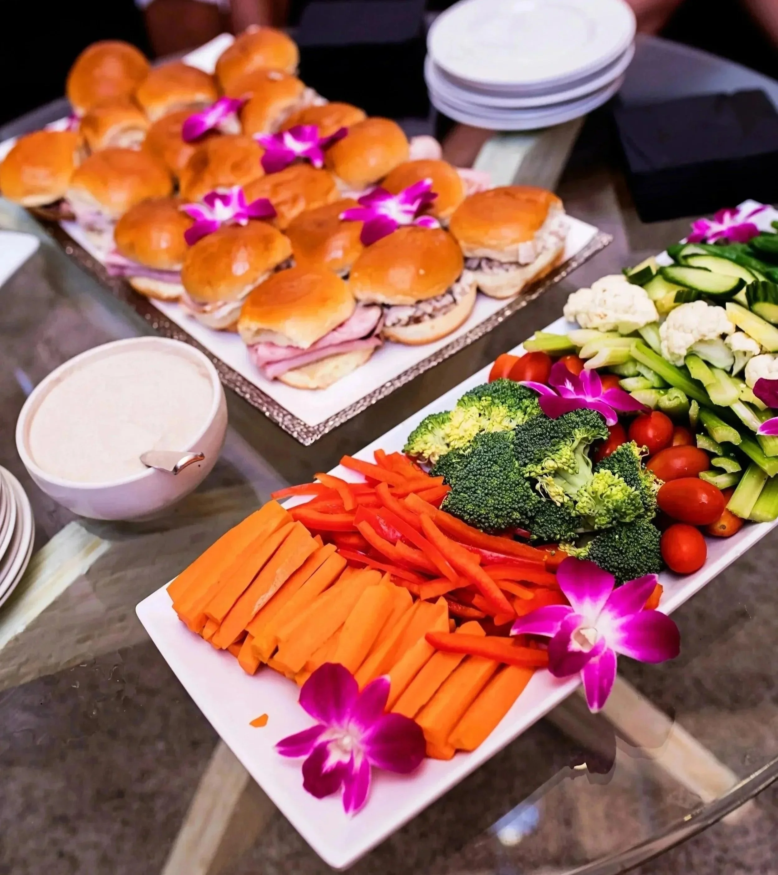 Tray of mini sandwiches with pink flowers on top, surrounded by bowls of dip and a large platter of fresh vegetables including sliced carrots, cherry tomatoes, broccoli, cauliflower, and cucumber at a buffet or party table.