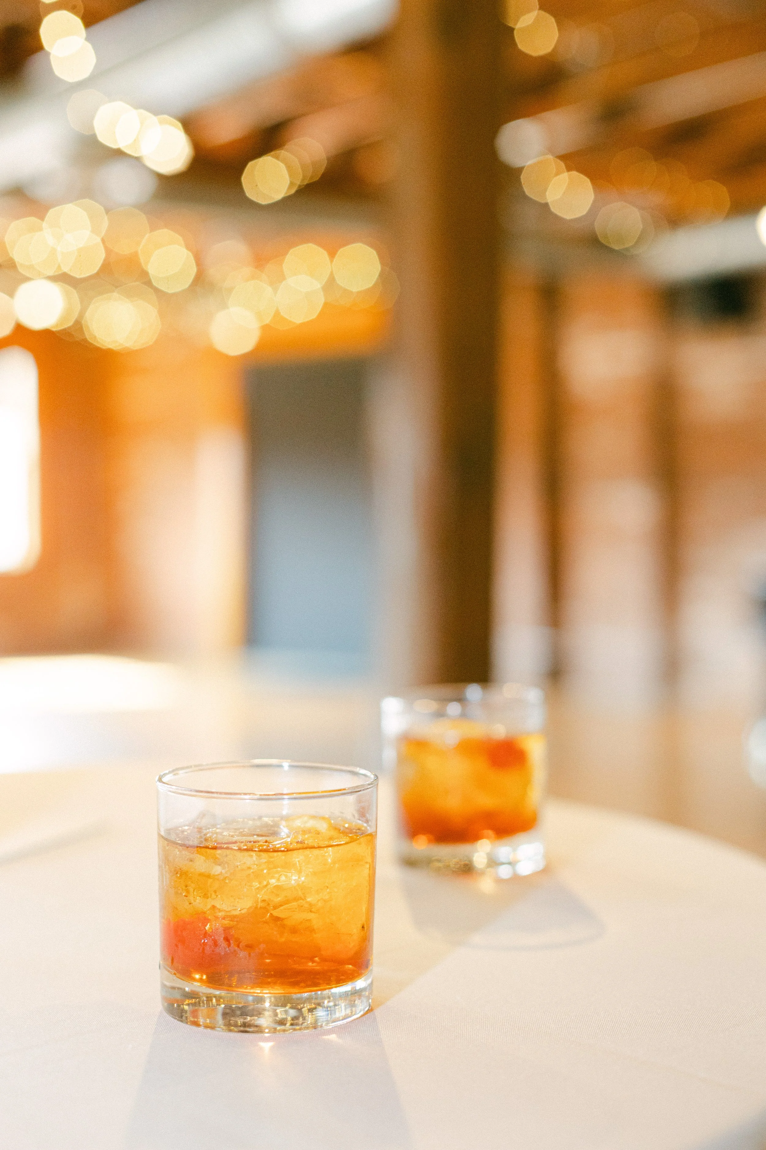 Two glasses of amber-colored drinks with ice cubes on a white surface in a warmly lit room.