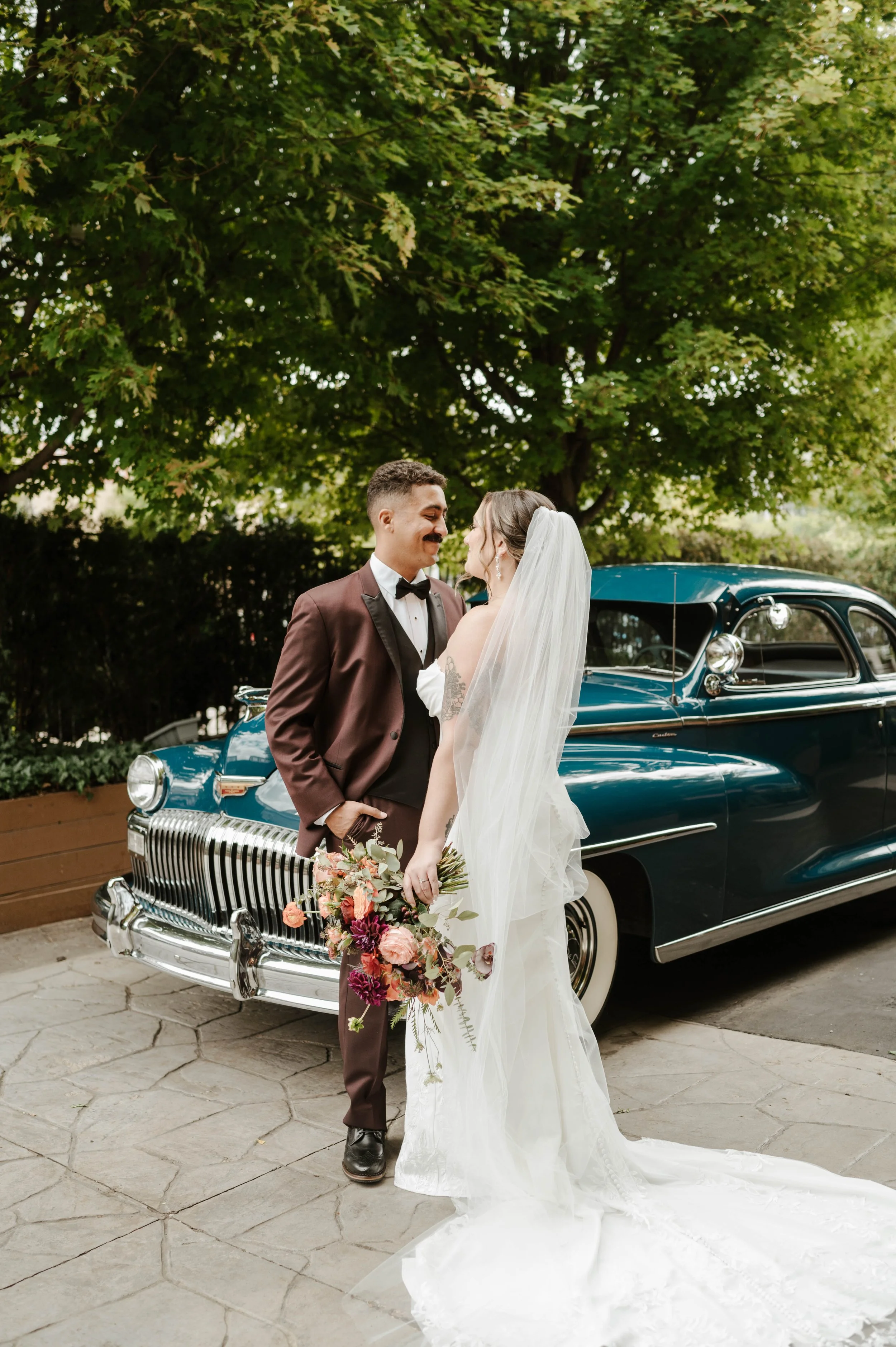 A bride and groom stand close together in front of a vintage blue car under a large green tree, with the bride holding a floral bouquet and wearing a long white wedding dress and veil, while the groom wears a brown tuxedo with a bow tie, smiling at e