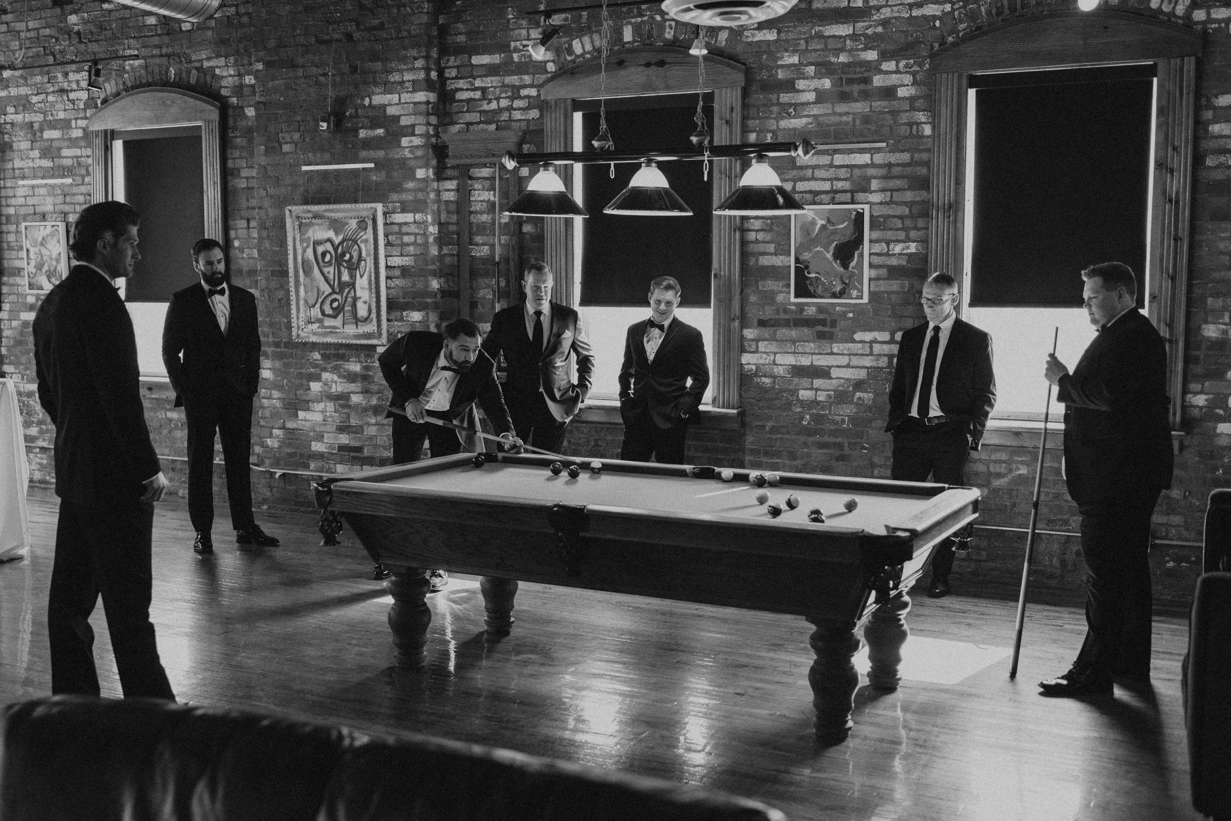 A group of men in tuxedos playing pool in a brick-walled room with artwork on the walls and windows in the background.
