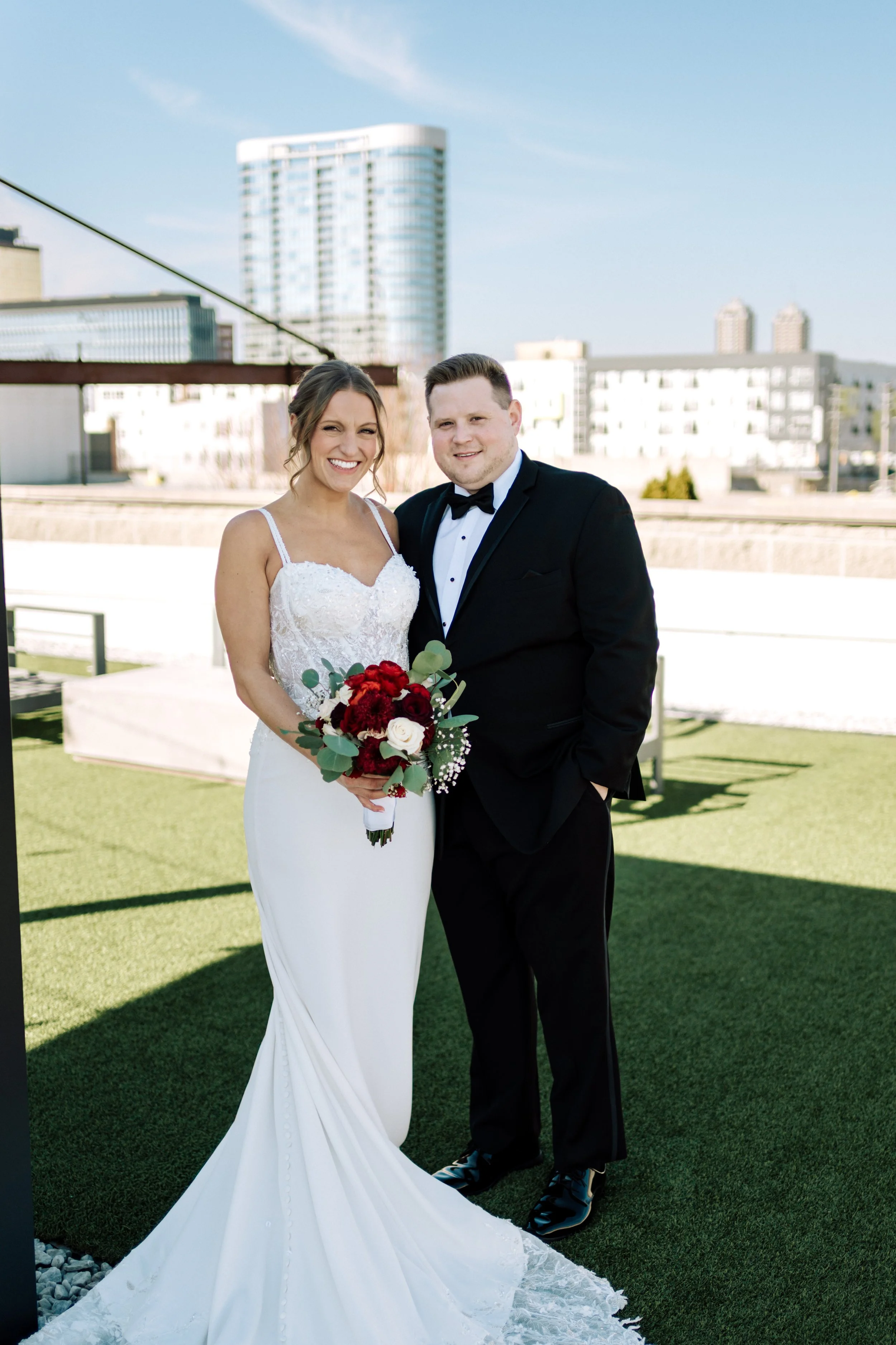 A bride and groom smiling together outdoors, the bride holding a bouquet of red and white flowers, with a city skyline in the background.