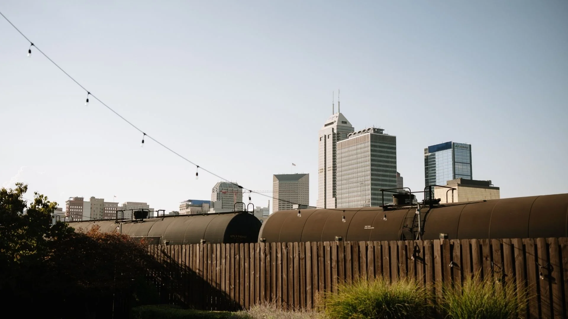 City skyline with tall modern buildings, string lights in the foreground, and a train or similar rail vehicle with a wooden fence and greenery in the lower part of the image.