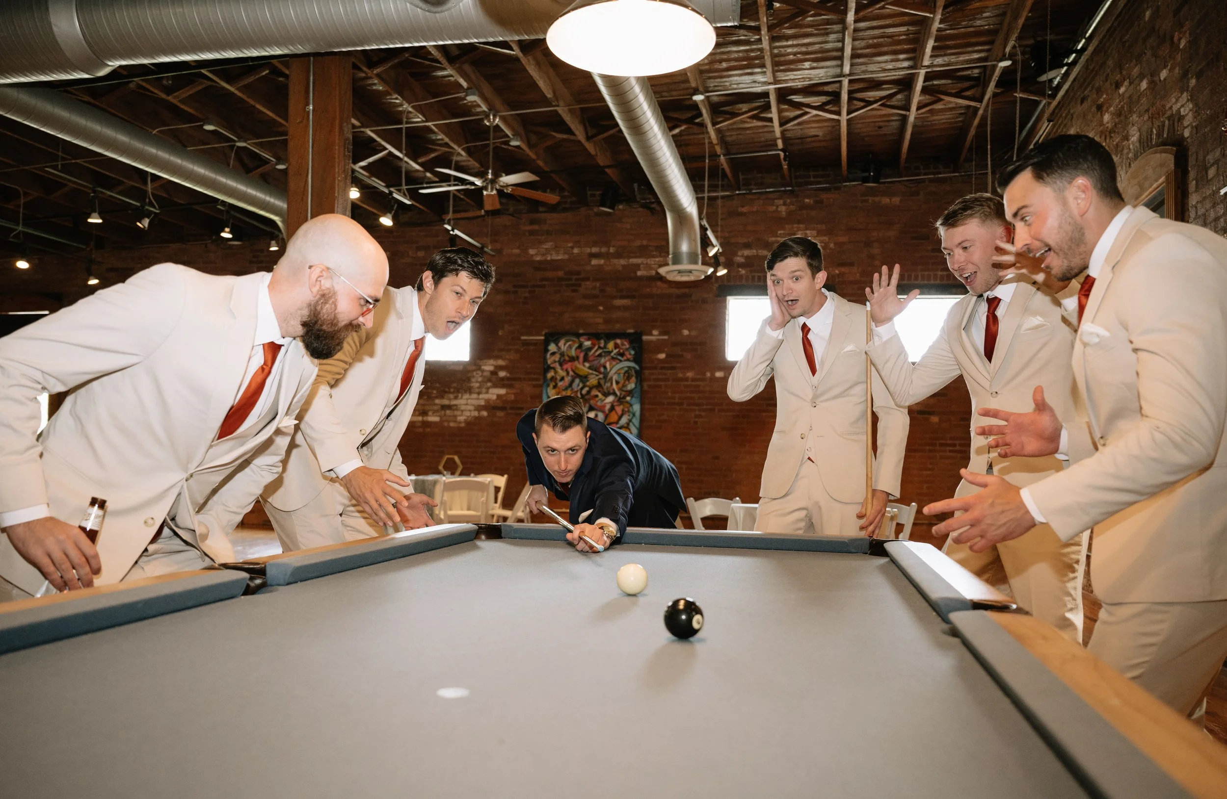 Group of men in suits playing pool in a rustic indoor setting with exposed brick walls and wooden beams.