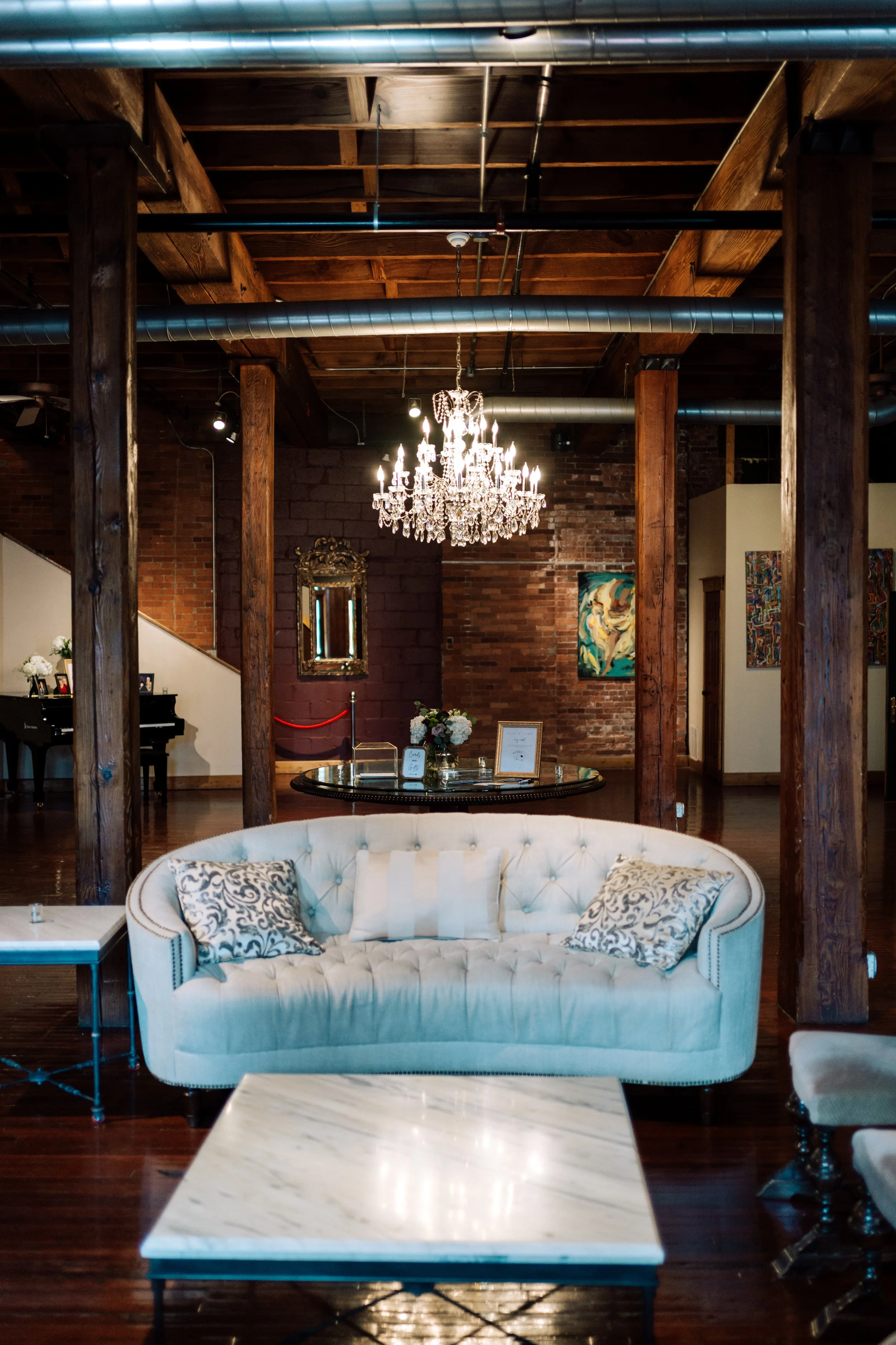The interior of a stylish room with exposed brick walls, wooden beams, a large crystal chandelier, a framed mirror, a colorful abstract painting, a black grand piano, and elegant furniture including a white tufted sofa with patterned pillows and a ma