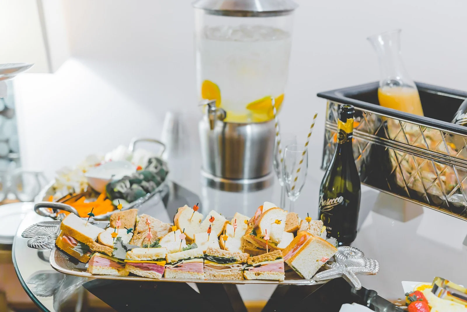 Assorted finger sandwiches, some with cold cuts, laid out on a silver platter, with a water dispenser with floating lemon slices, a bottle of sparkling beverage, and a tray with a glass container of orange juice in the background.