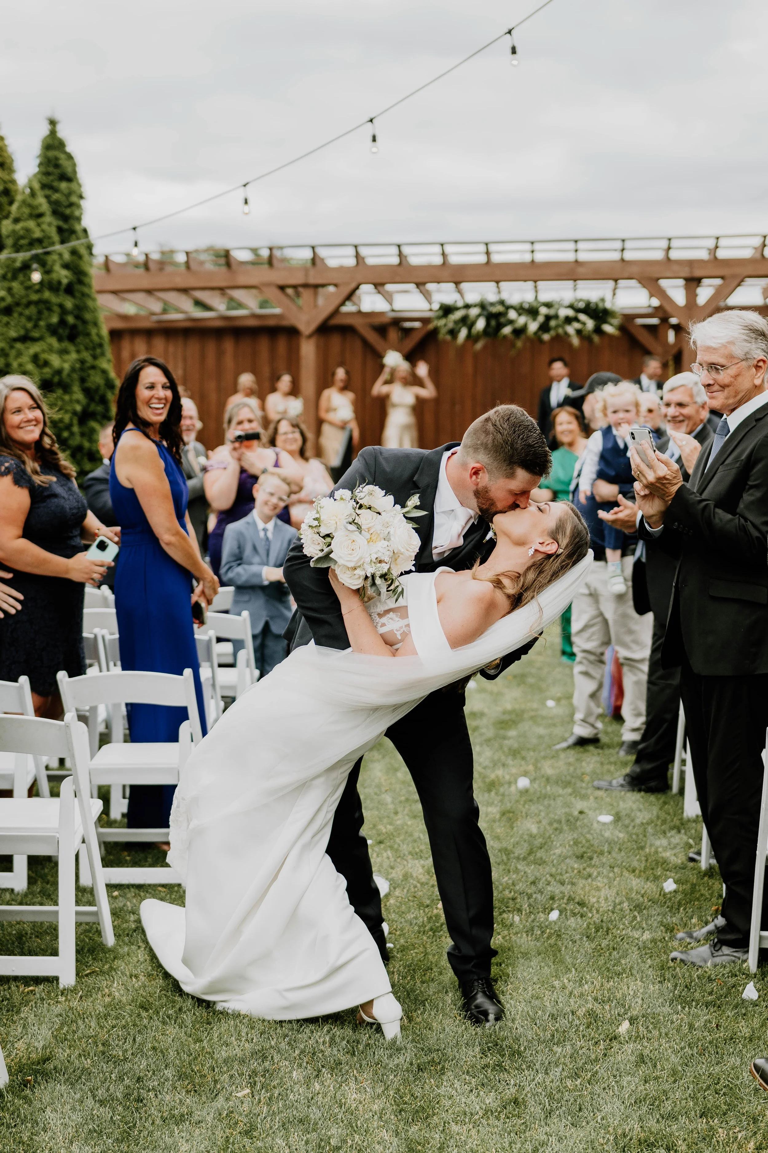 A bride and groom sharing a kiss during their outdoor wedding ceremony, with guests smiling and taking pictures in the background.