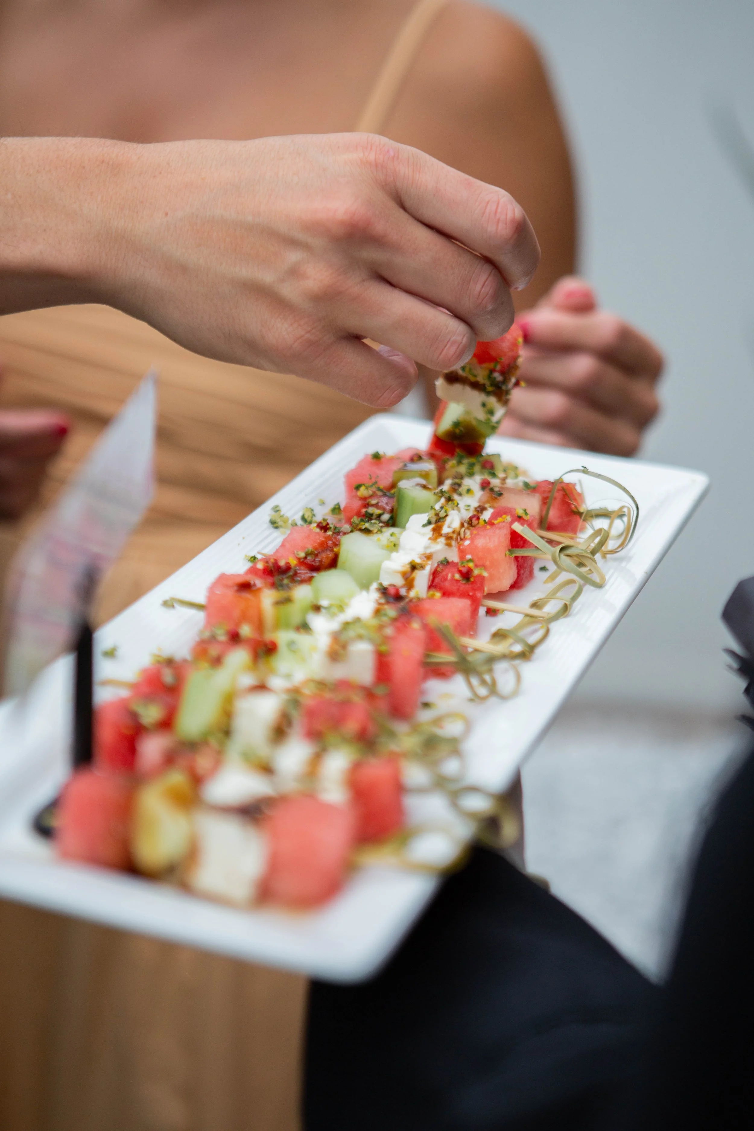 Person holding a square plate of watermelon, cucumber, and cheese salad garnished with herbs.