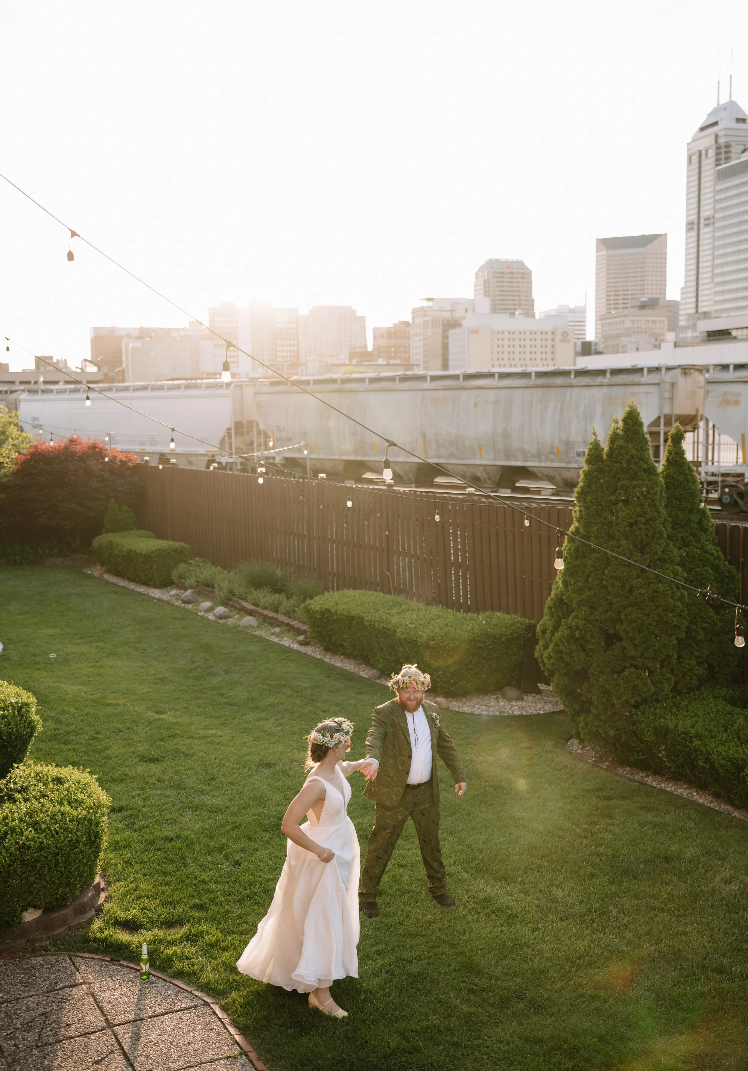 A couple dressed in wedding attire holding hands and walking in a lush green garden, with an urban city skyline in the background during sunset, and string lights overhead.