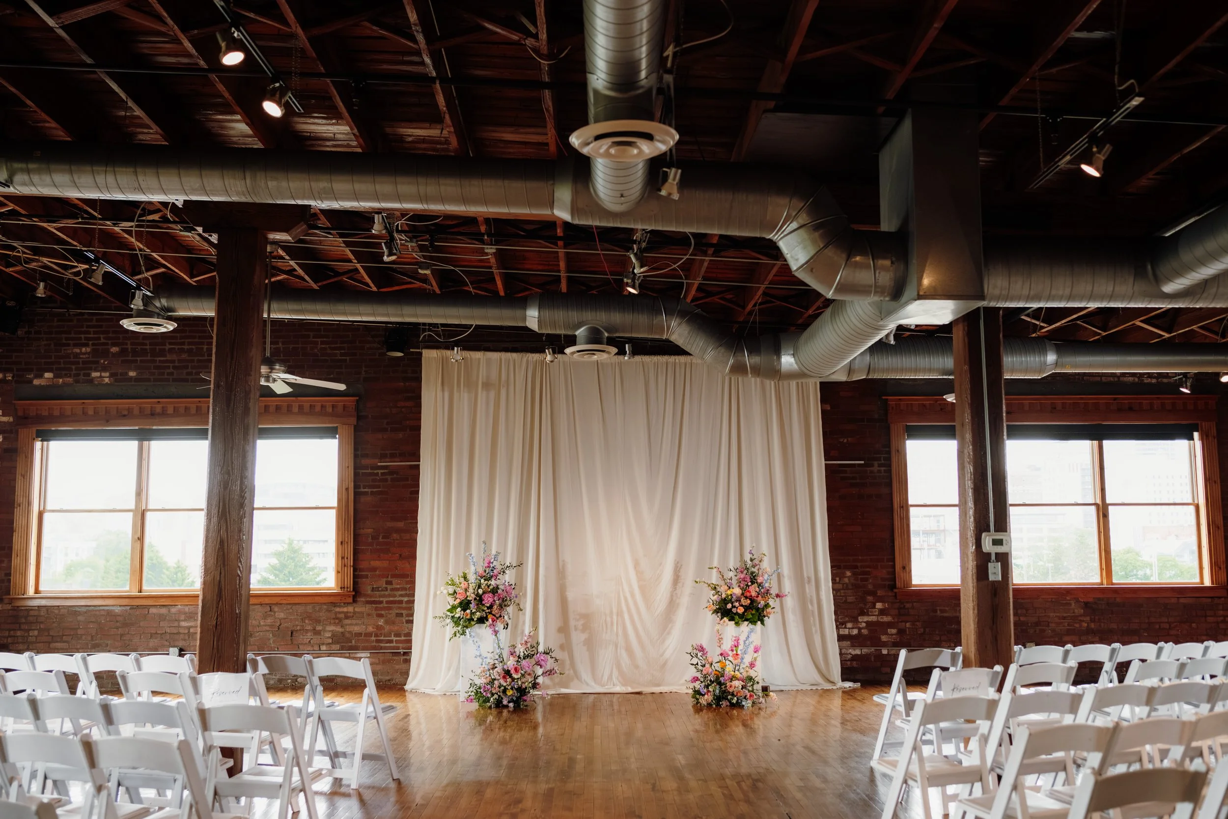 Indoor wedding venue with wooden floors, brick walls, large windows, rows of white chairs, floral arrangements, and a draped backdrop for a ceremony.