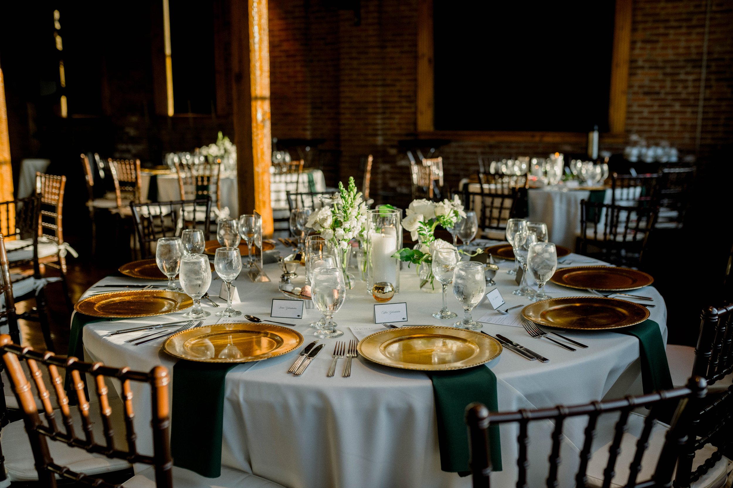 A round banquet table set for a formal event with white tablecloths, gold charger plates, wine glasses, water glasses, silverware, and floral centerpieces in glass vases. The background shows a rustic interior with brick walls and wooden beams.