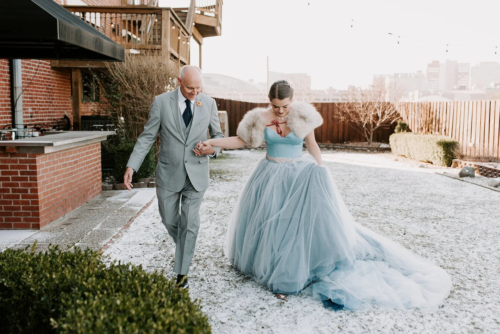 A bride in a light blue gown and fur stole walking in a snowy backyard with an elderly man in a gray suit, holding hands and smiling.