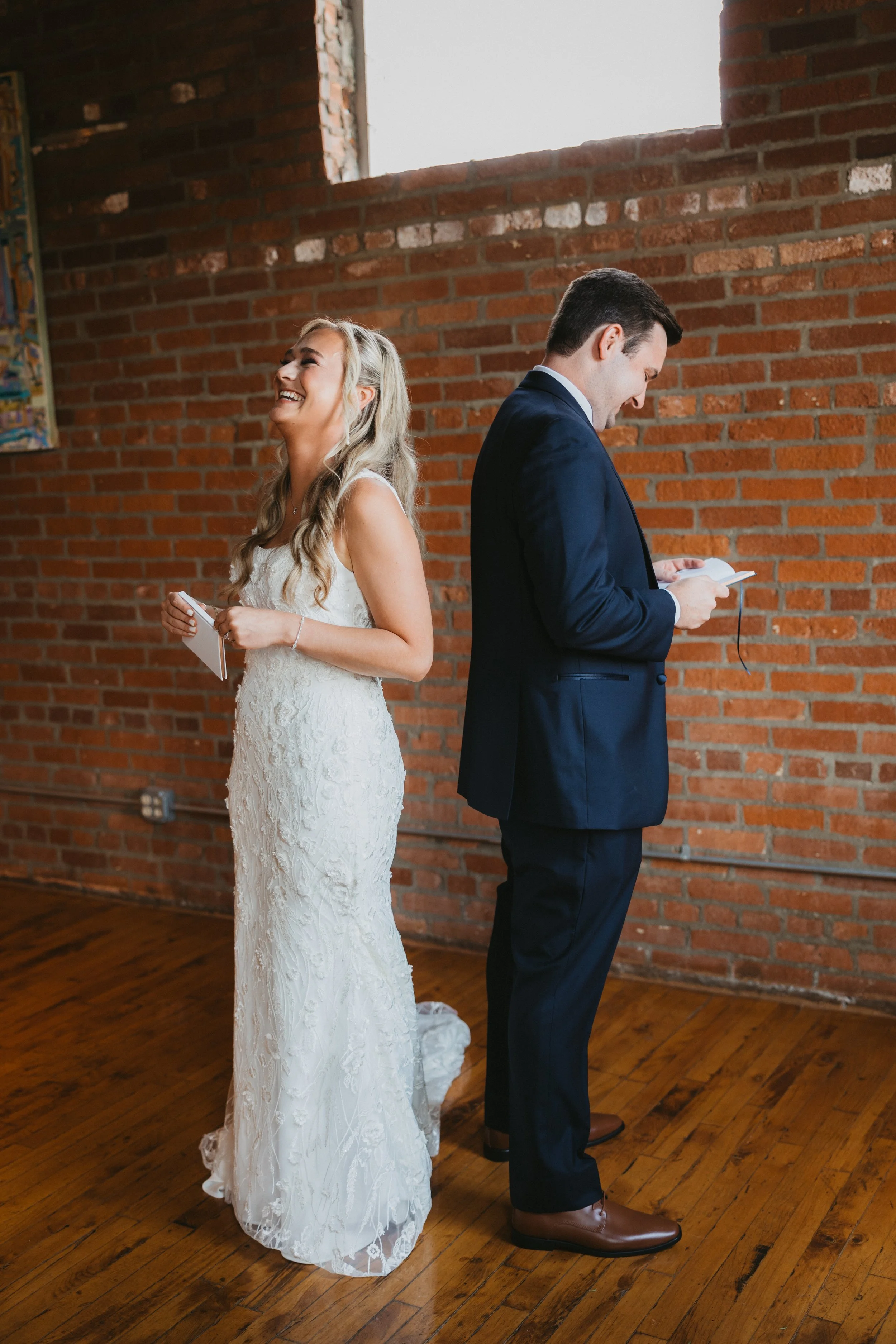 A bride and groom stand back-to-back inside a room with a brick wall, holding small notebooks. The bride is laughing and dressed in a white lace wedding gown, while the groom is looking down at his notebook and dressed in a dark suit with brown shoes