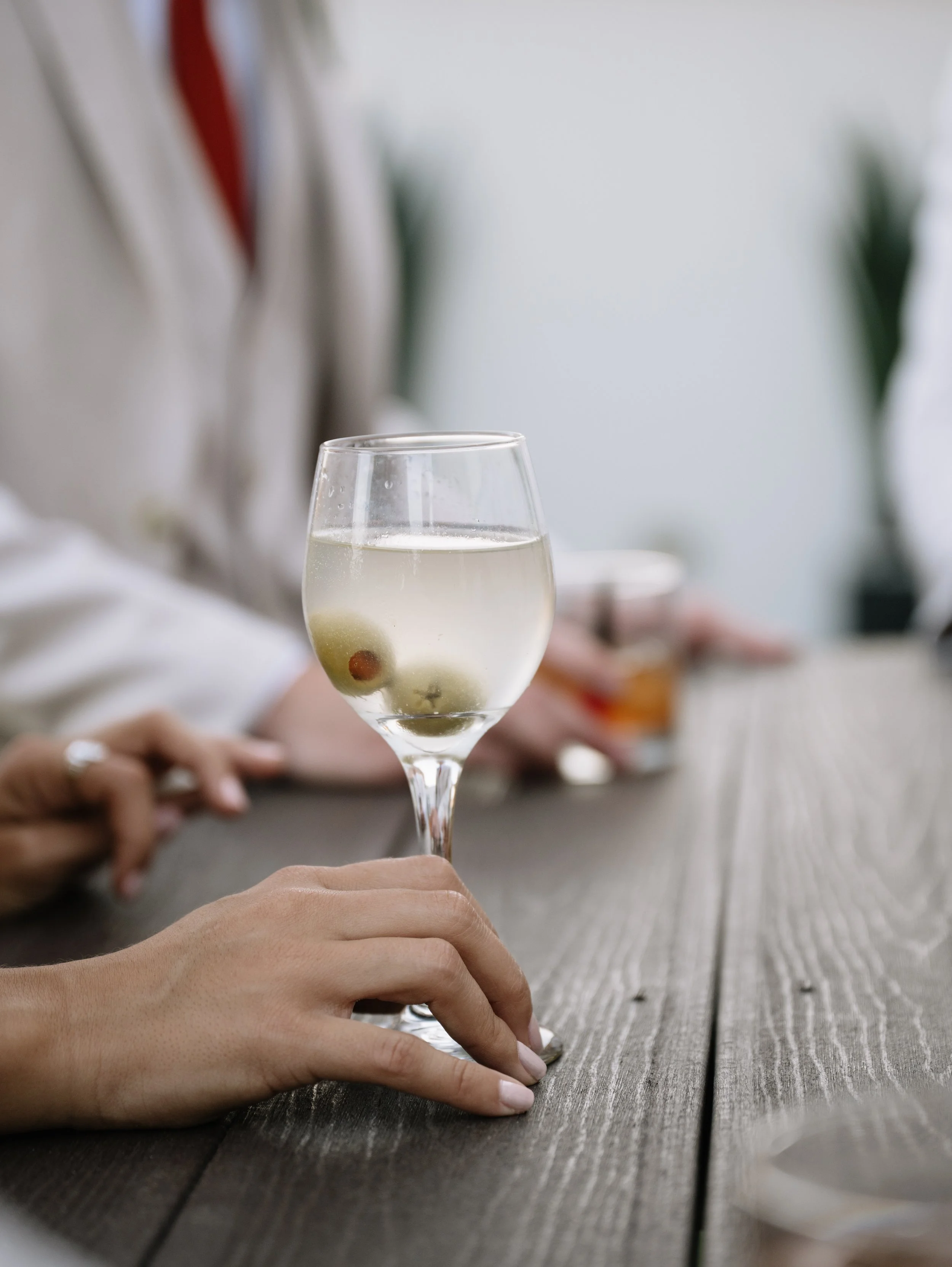 A person holding a glass of white wine with olives inside, sitting at a wooden table in a social setting.