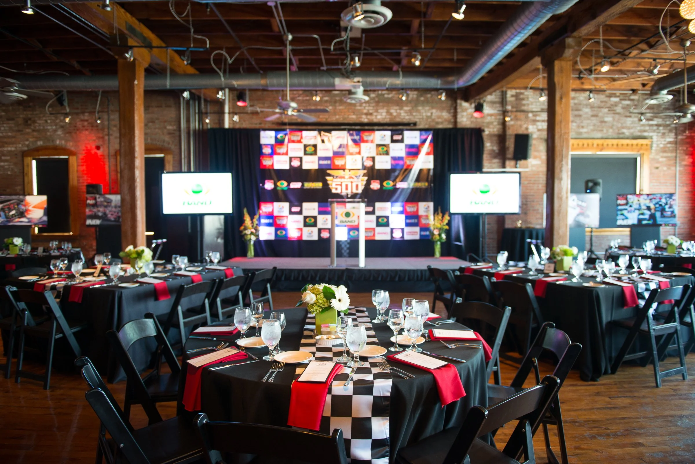 Indoor event space set up for a race-themed gathering with tables decorated in black, white, and red, featuring checkered tablecloths and floral centerpieces. A stage with a black curtain backdrop displays a large checkered logo and various sponsor b