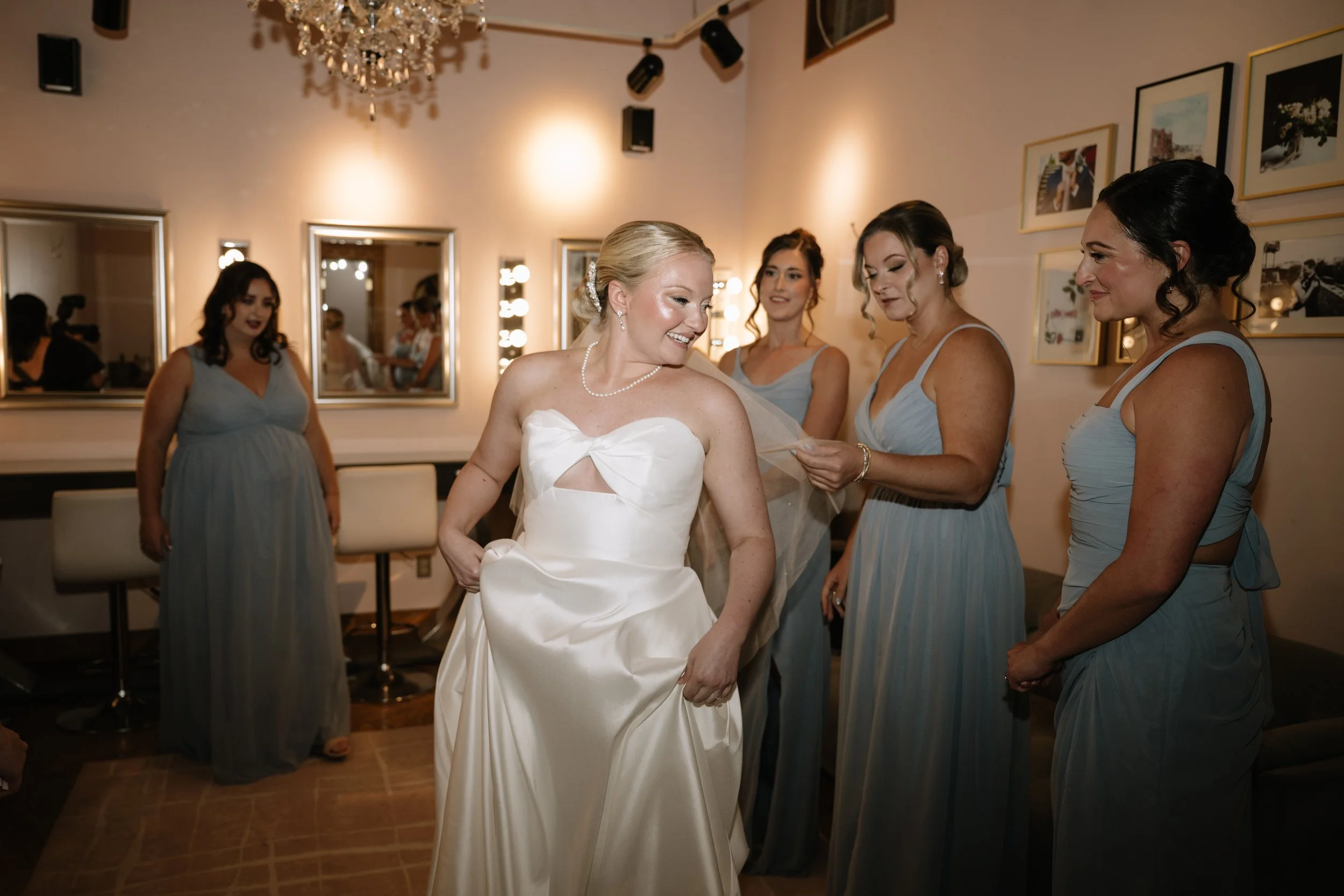A bride in a white wedding dress smiling as she stands with her bridesmaids in light blue dresses in a decorated room.