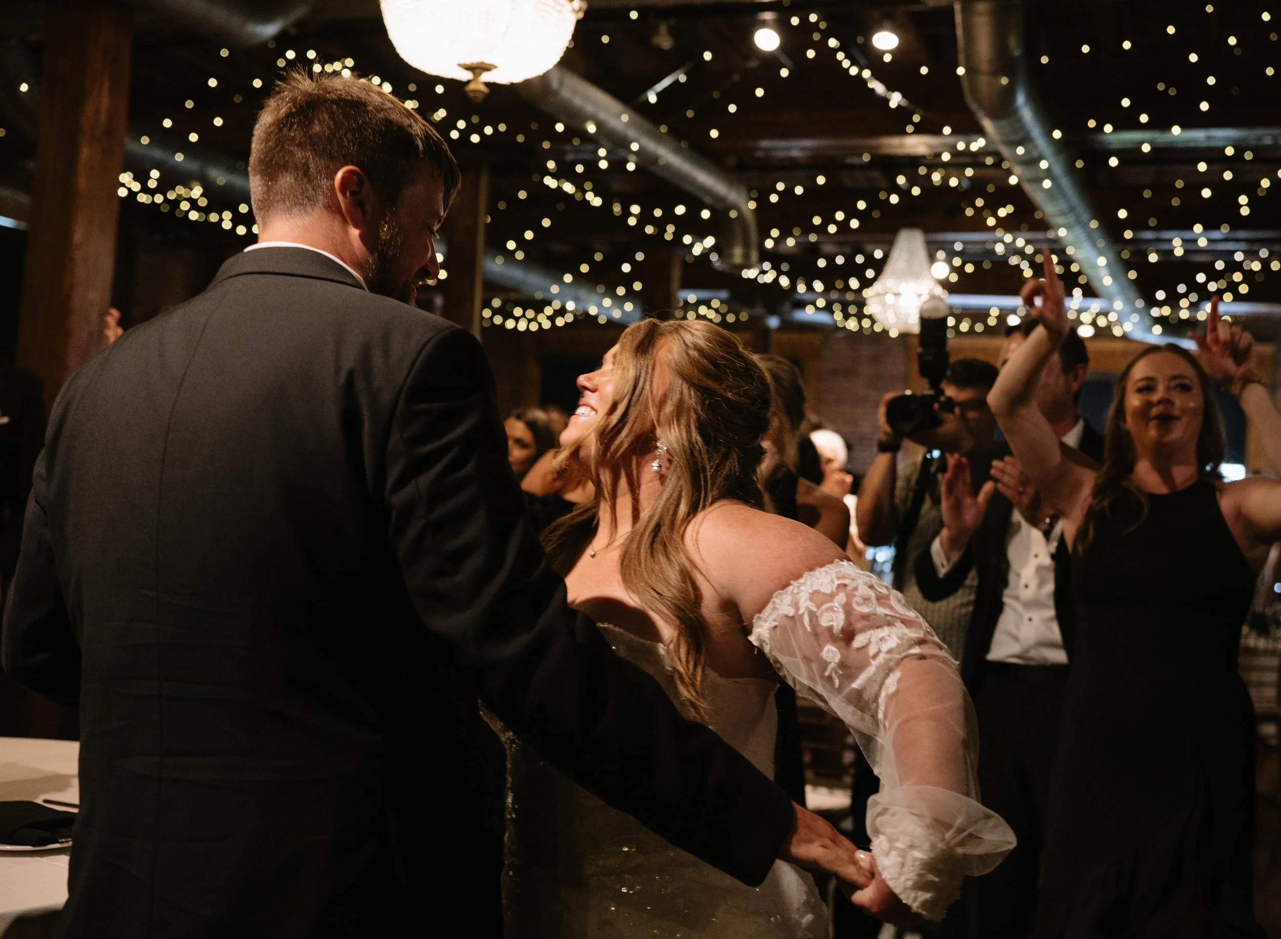 A bride and groom share a kiss during their wedding reception, surrounded by guests under string lights and chandeliers in a rustic, indoor venue.