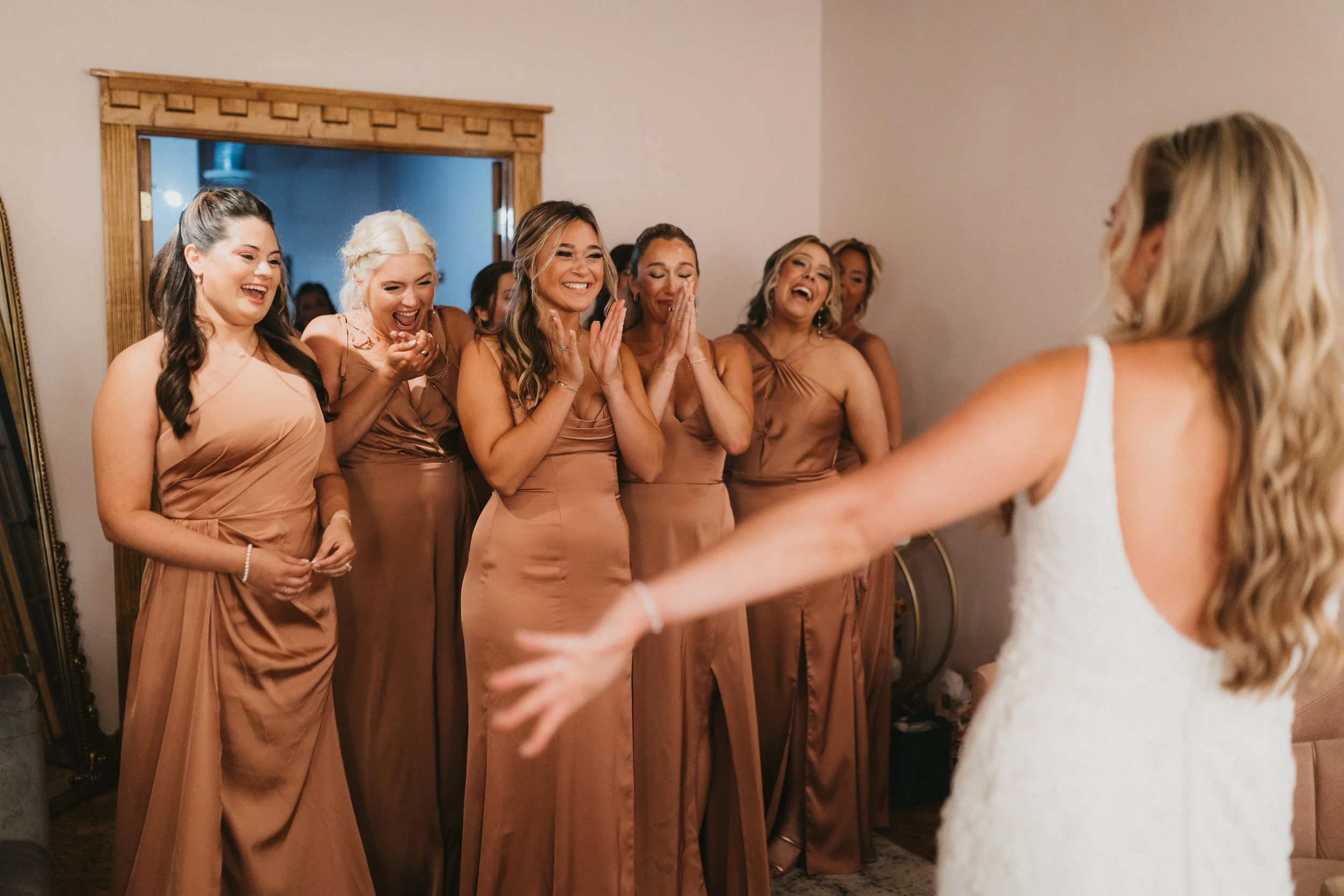A group of women dressed in matching champagne-colored dresses appear to be watching a bride in a white wedding dress as she walks into the room, all of them smiling and laughing with joy.
