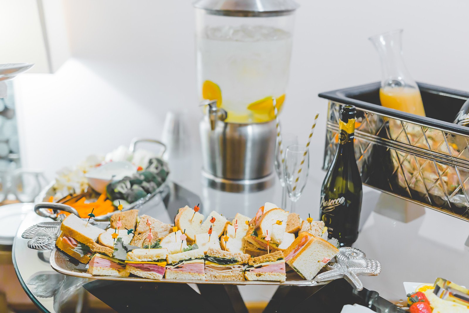 Assorted finger sandwiches on a silver tray with toothpicks, including ham and cheese, egg salad, and cucumber sandwiches, part of a buffet spread with a beverage dispenser, a champagne bottle, and a beverage jug in the background.