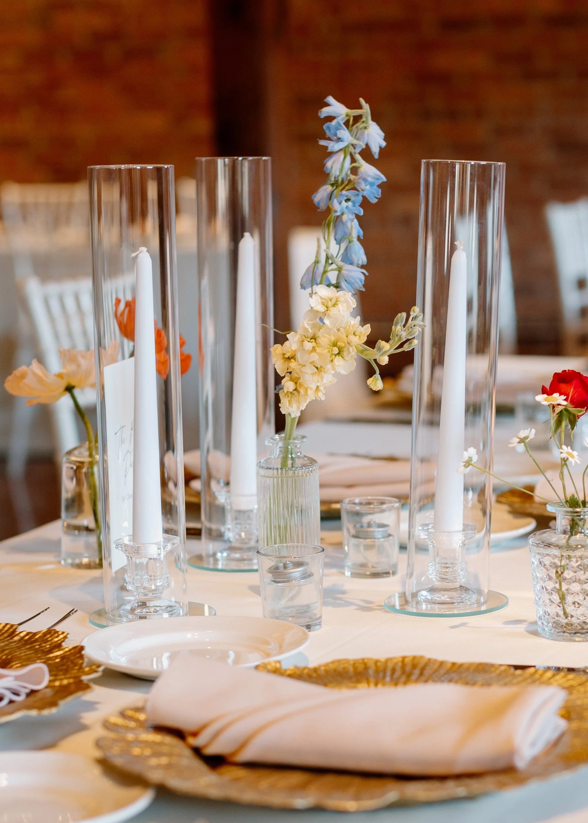 A decorated dining table with tall glass candle holders, white candles, and floral arrangements, including light blue and white flowers, in a restaurant or event setting with brick walls in the background.